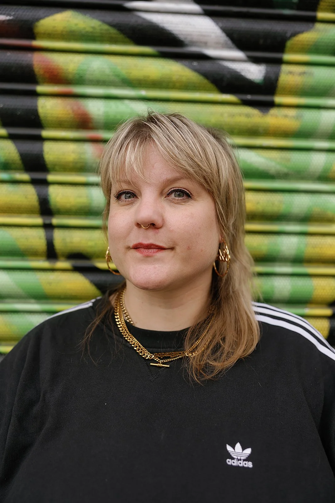 A woman with blonde hair and light skin posing in front of a colorful wall with graffiti.