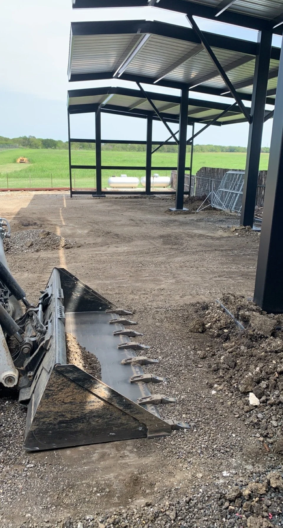 Construction site with a partially built metal structure and a bulldozer attachment ground. Green fields and propane tanks in the background.