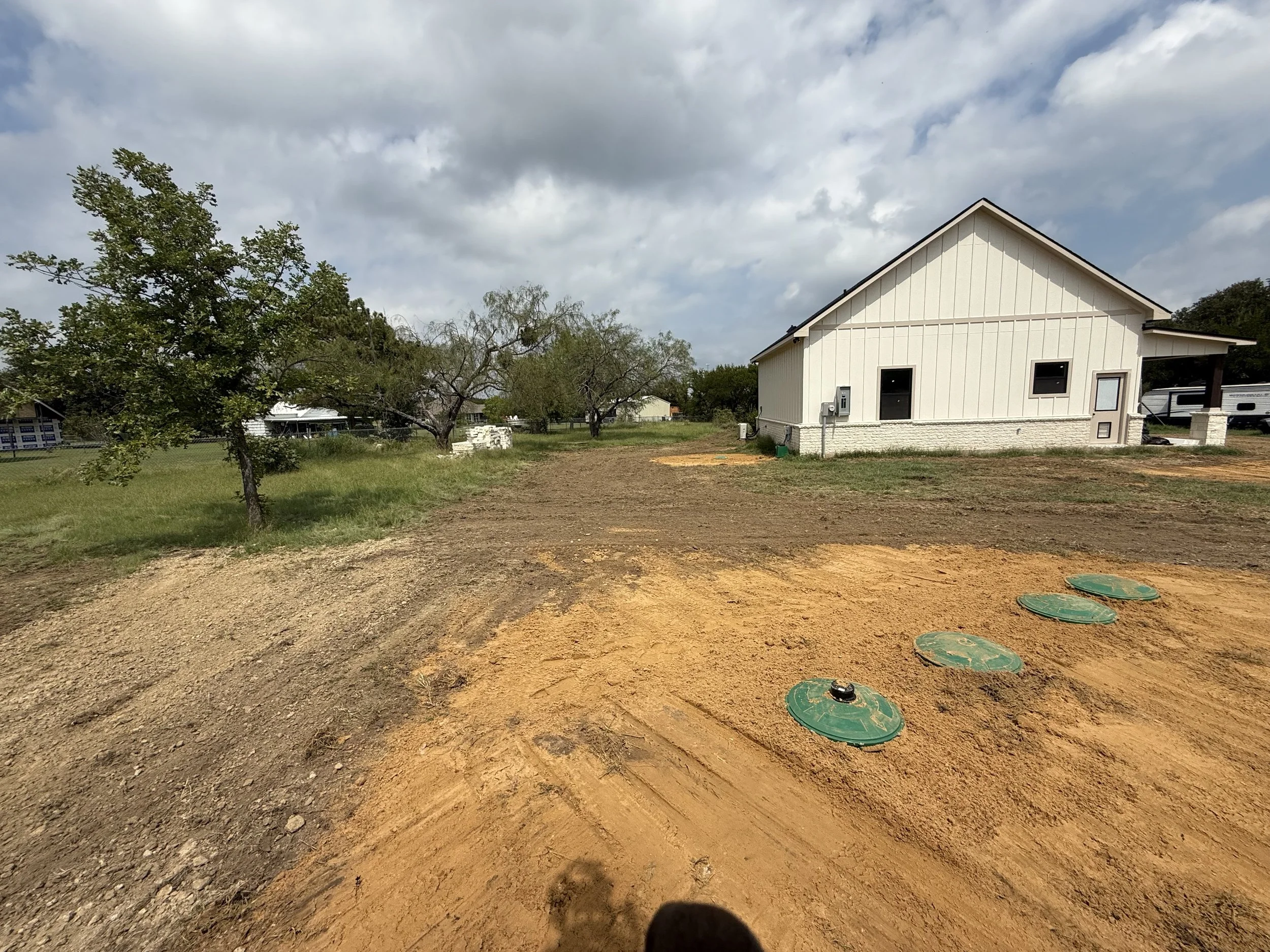 Residential house under construction with a dirt yard and utility access covers in the foreground, trees, and a cloudy sky in the background.