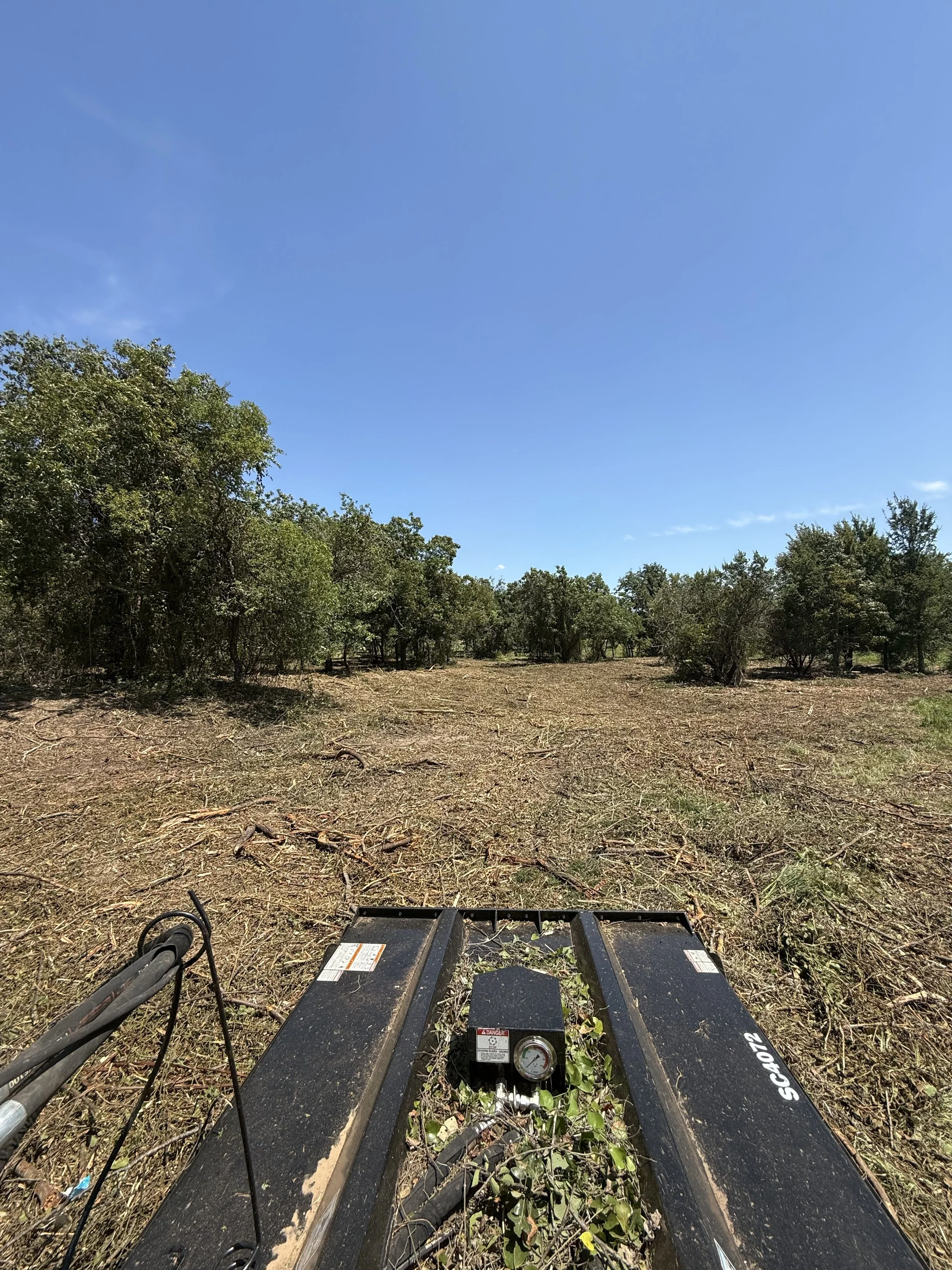 View from a land clearing with a black brush cutter in the foreground, partly overgrown with weeds, and trees on the horizon under a blue sky.