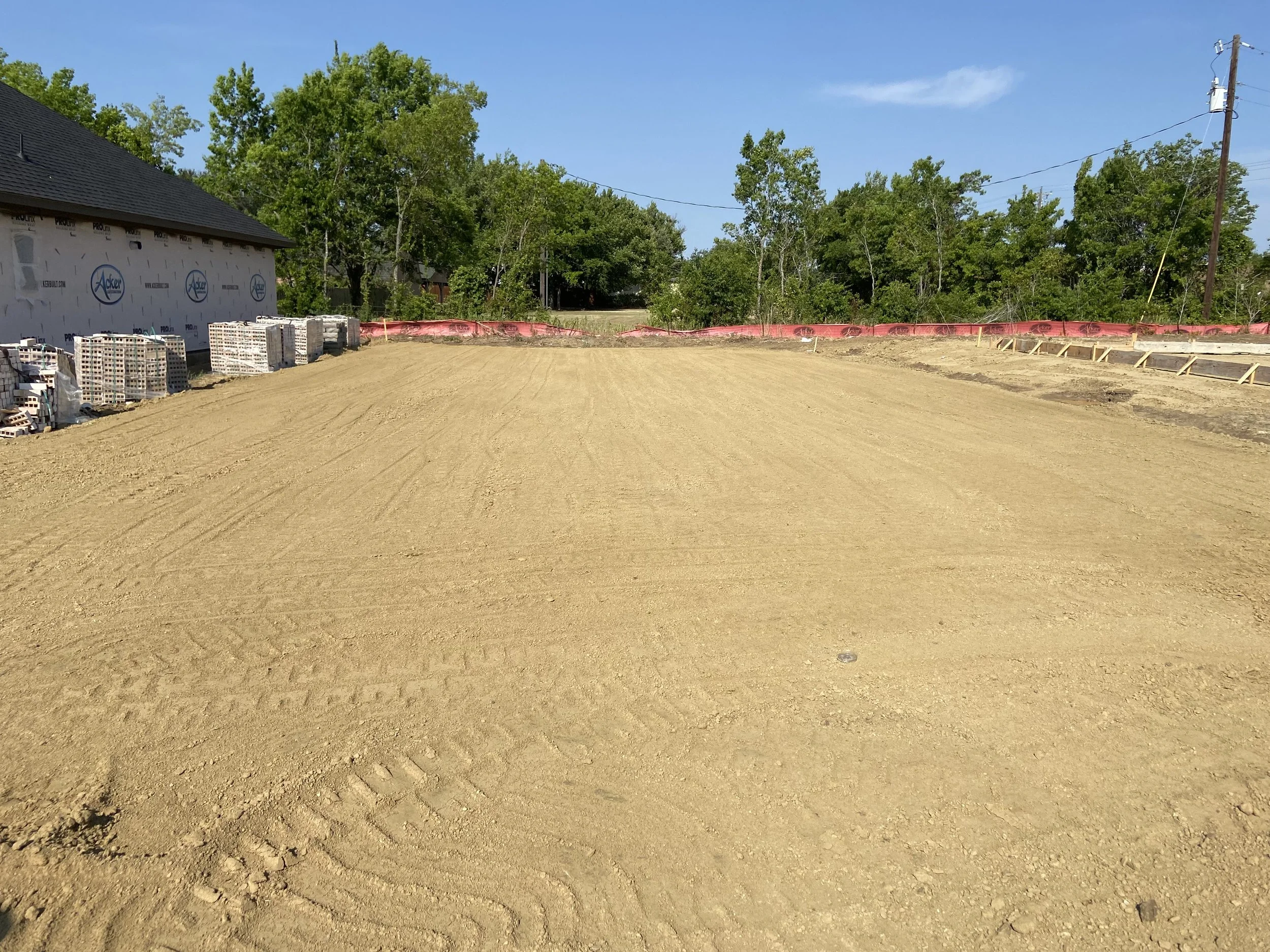 A construction site with a cleared dirt area, some stacks of bricks or blocks on the left, and a partially built structure on the left side of the image. Green trees and a blue sky with some clouds are visible in the background.