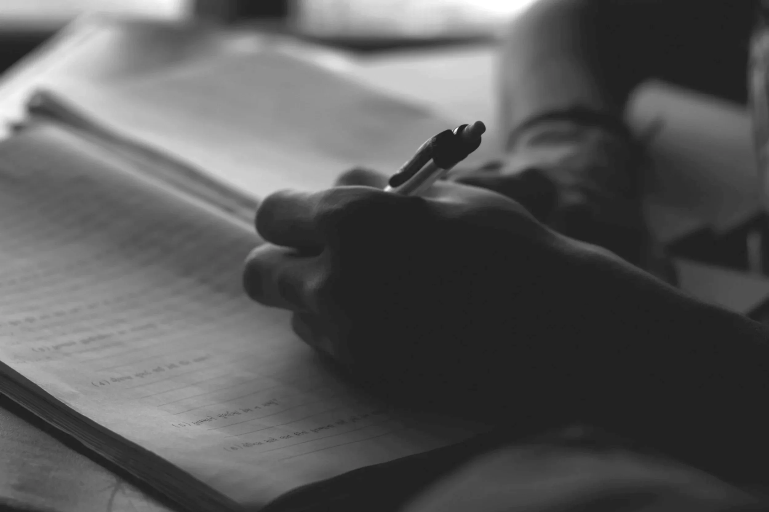 Close-up of a person's hand holding a pen and writing in a notebook on a desk.