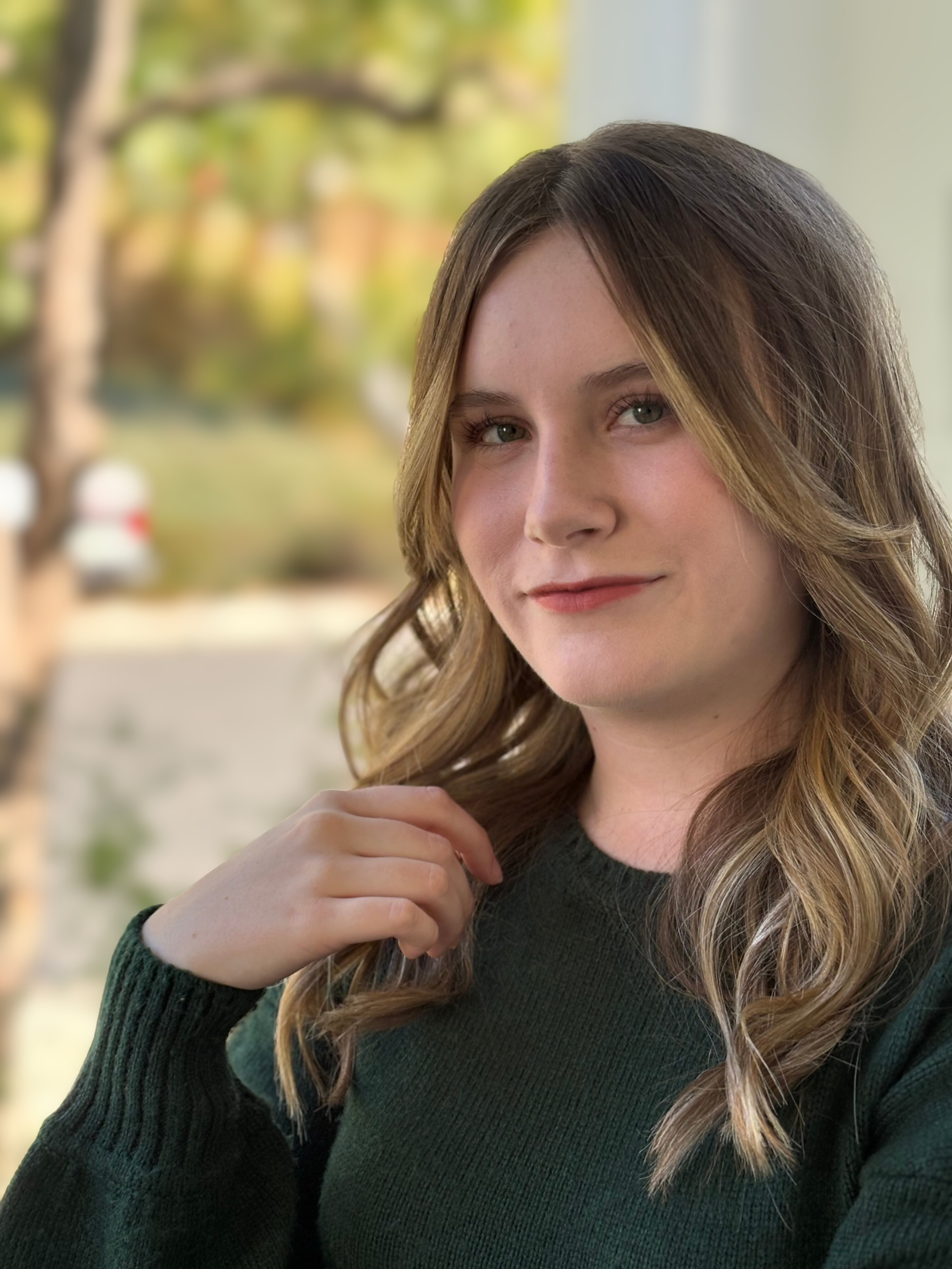 Portrait of a young woman with wavy brown hair and green eyes, wearing a dark green sweater, outdoors with blurred trees in the background.