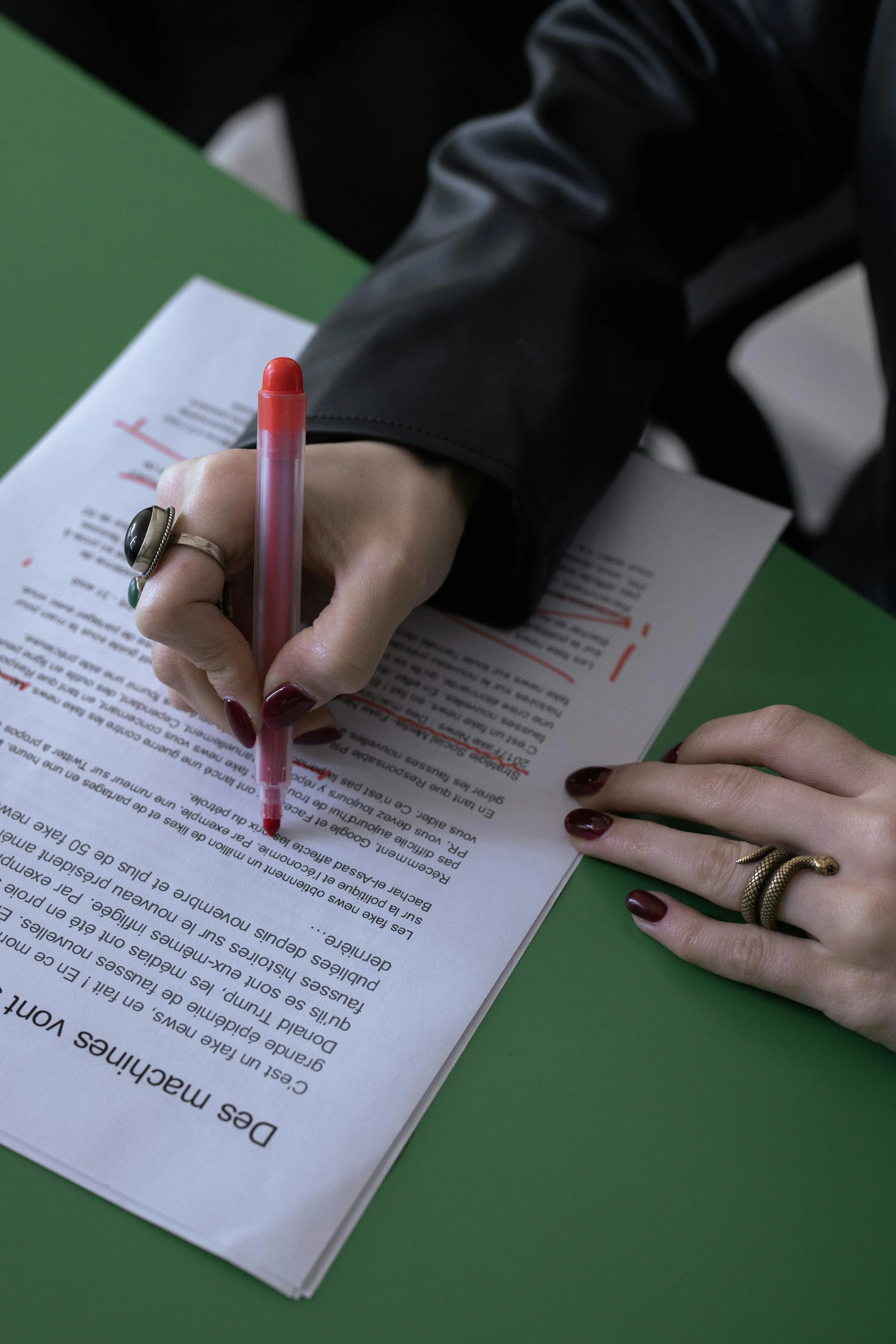 Close-up of hands, one with rings, signing a document with red pen on green table.