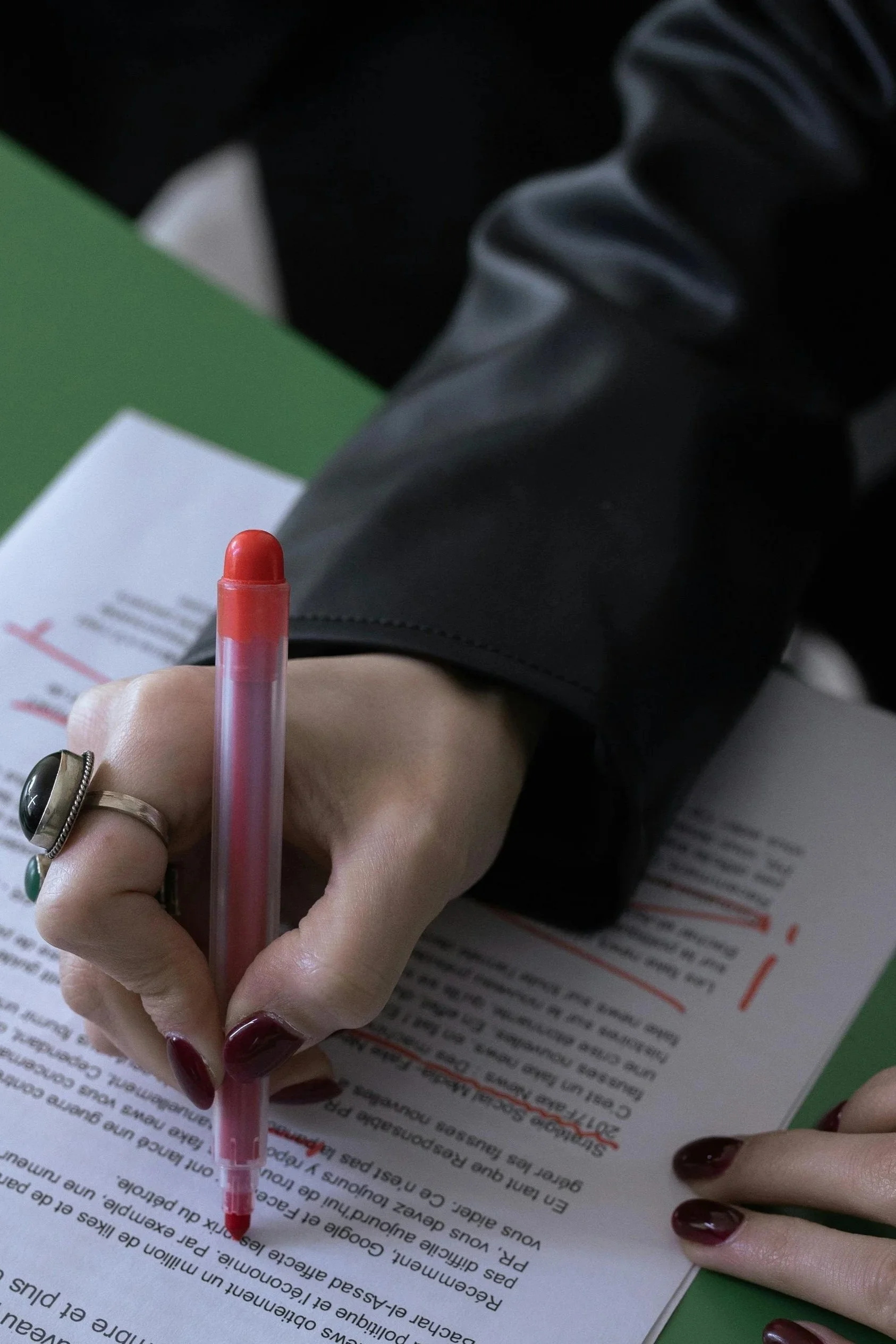 Close-up of a person's hand holding a pink pen, writing on a document with red and black text, with a black jacket sleeve, green paper folder, and printed paper in the background.