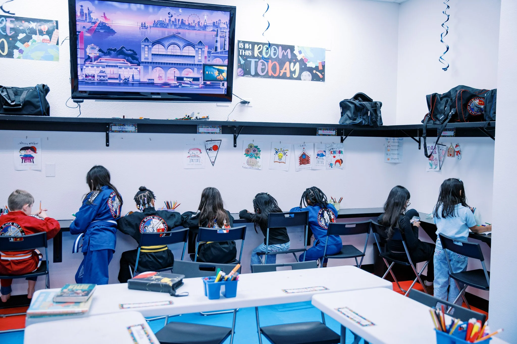 Children in a classroom sitting at a counter, drawing with colored pencils. The classroom has children's artwork displayed on the wall and a large television screen mounted on the wall showing a cityscape.