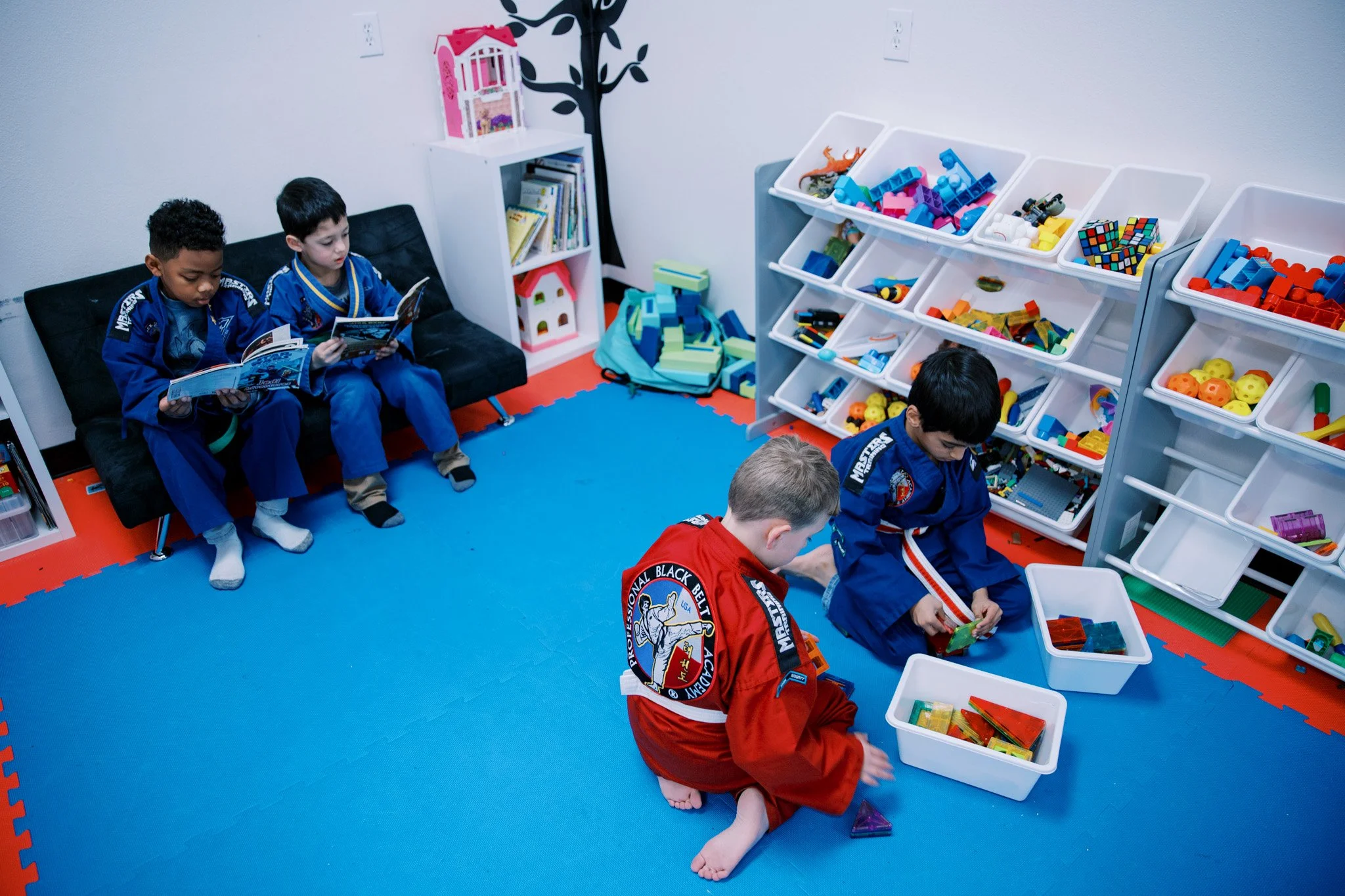 Four children in martial arts uniforms playing with building blocks in a playroom with toy storage shelves and books.