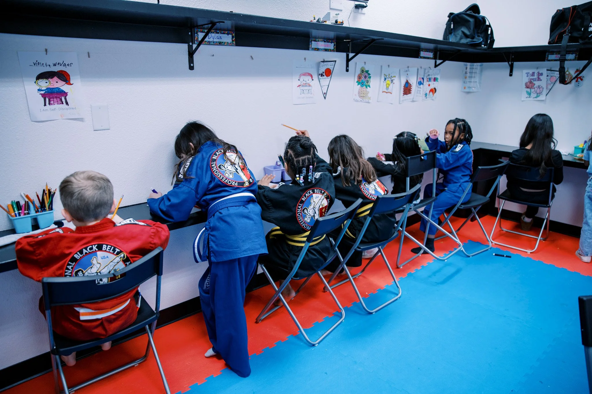 Children in martial arts uniforms sitting at desks, drawing or coloring in a classroom with martial arts posters on the wall.
