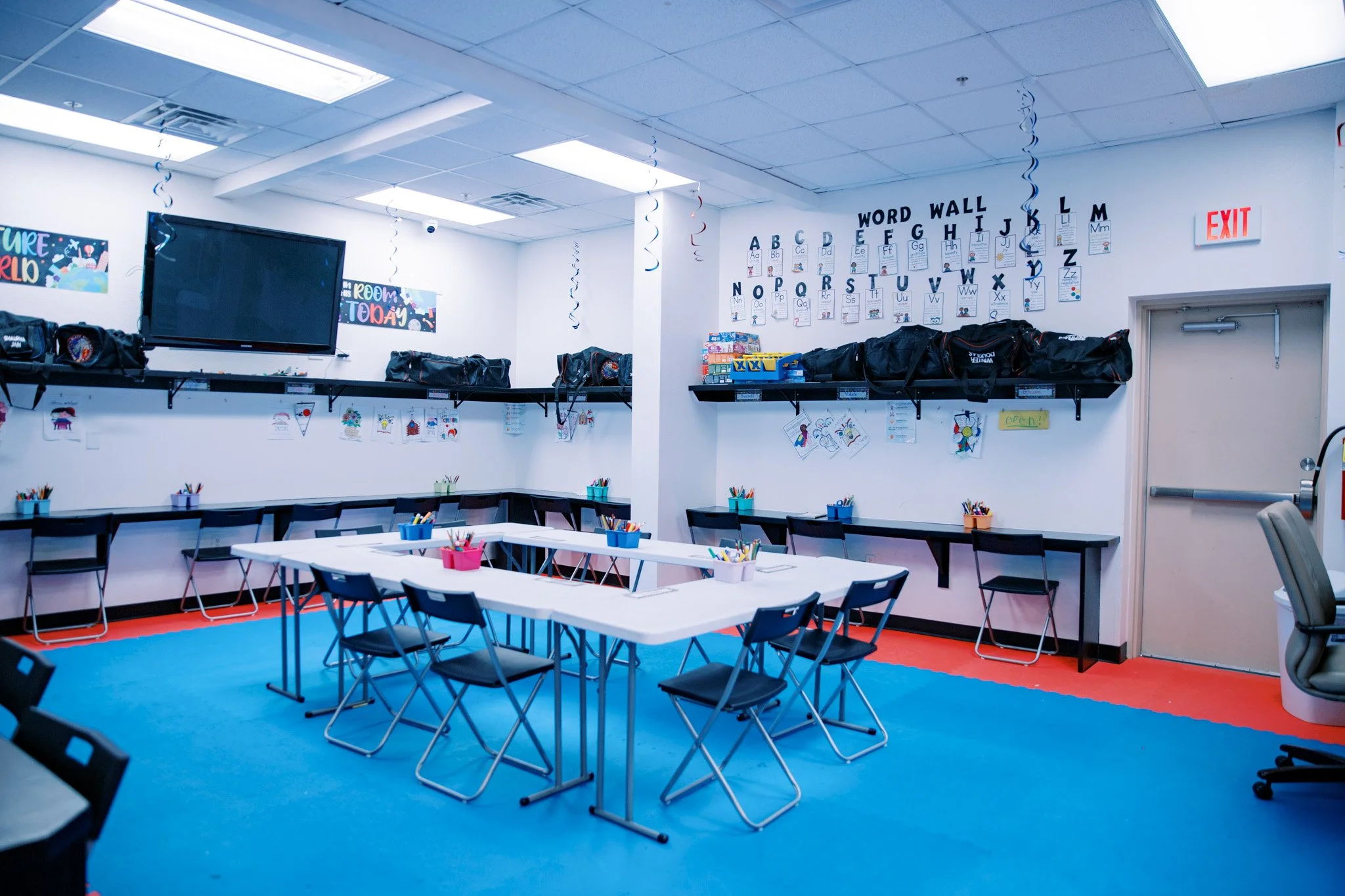 Classroom with tables and chairs, decorated for young students, featuring a word wall with alphabet letters, artwork, and storage bags along the wall.