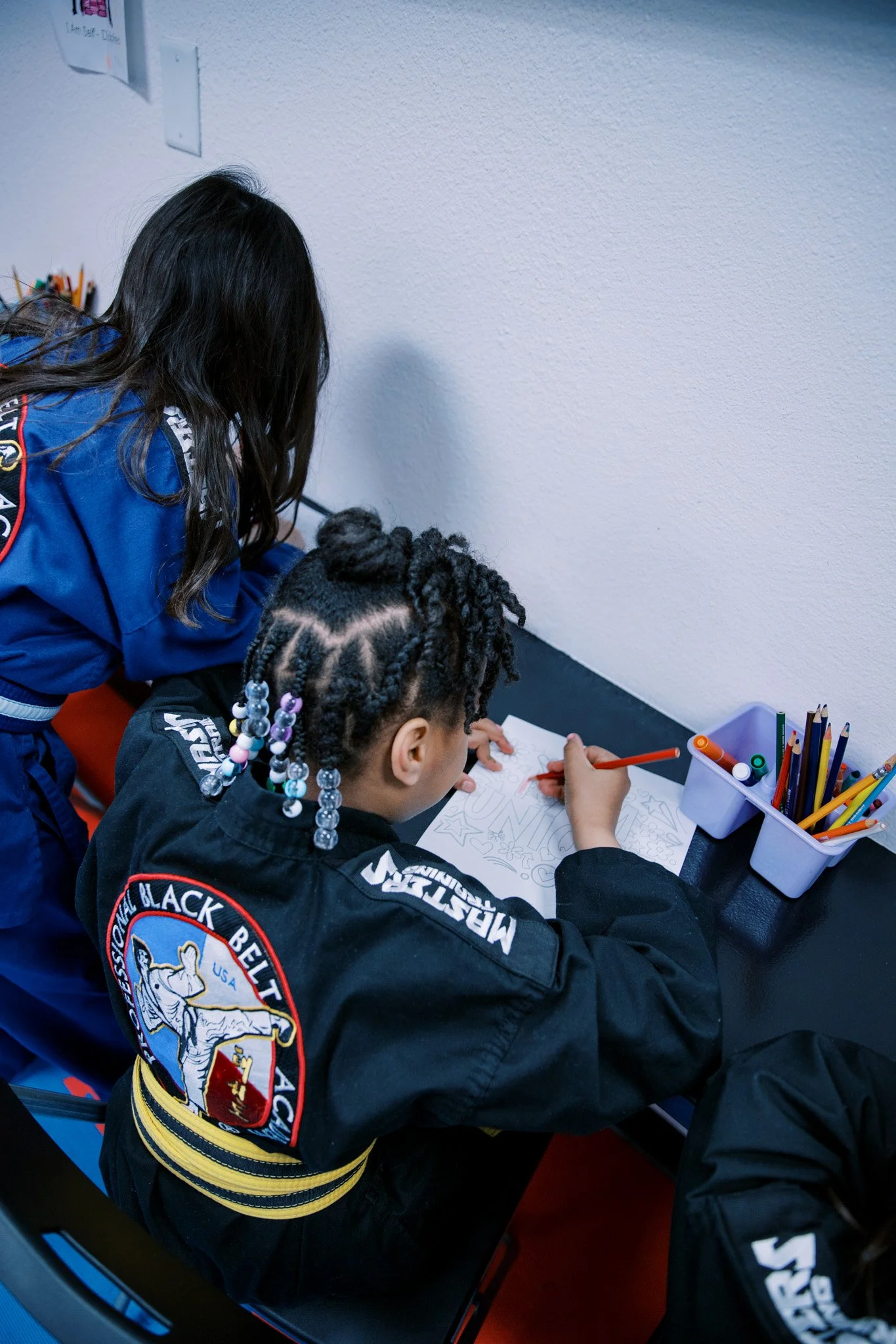 Two young girls in martial arts uniforms, black and blue, are working on coloring pages at a desk with colored pencils.