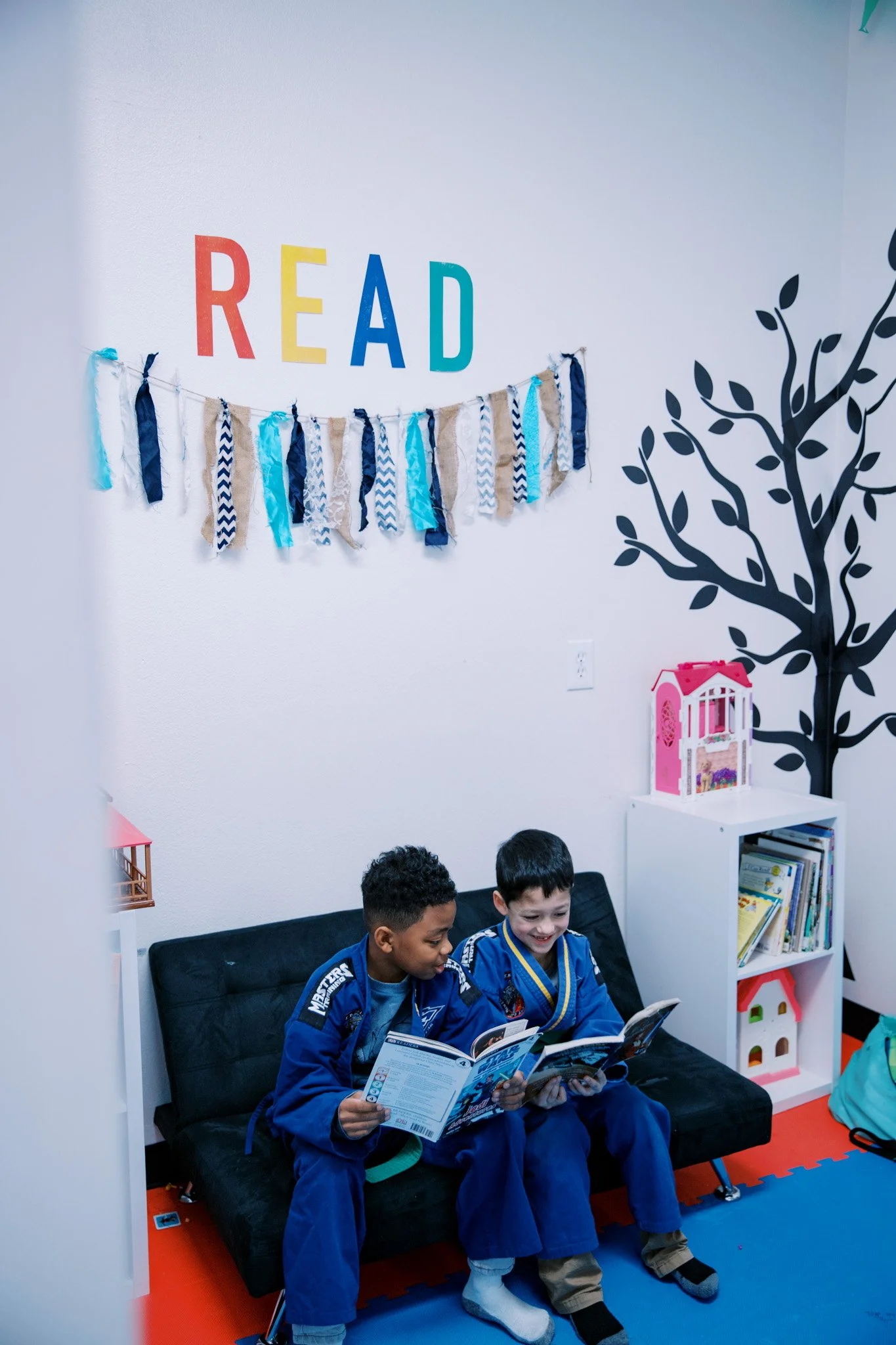 Two boys in martial arts uniforms reading books on a black couch in a colorful, decorated room, with the word 'READ' on the wall and a tree mural.