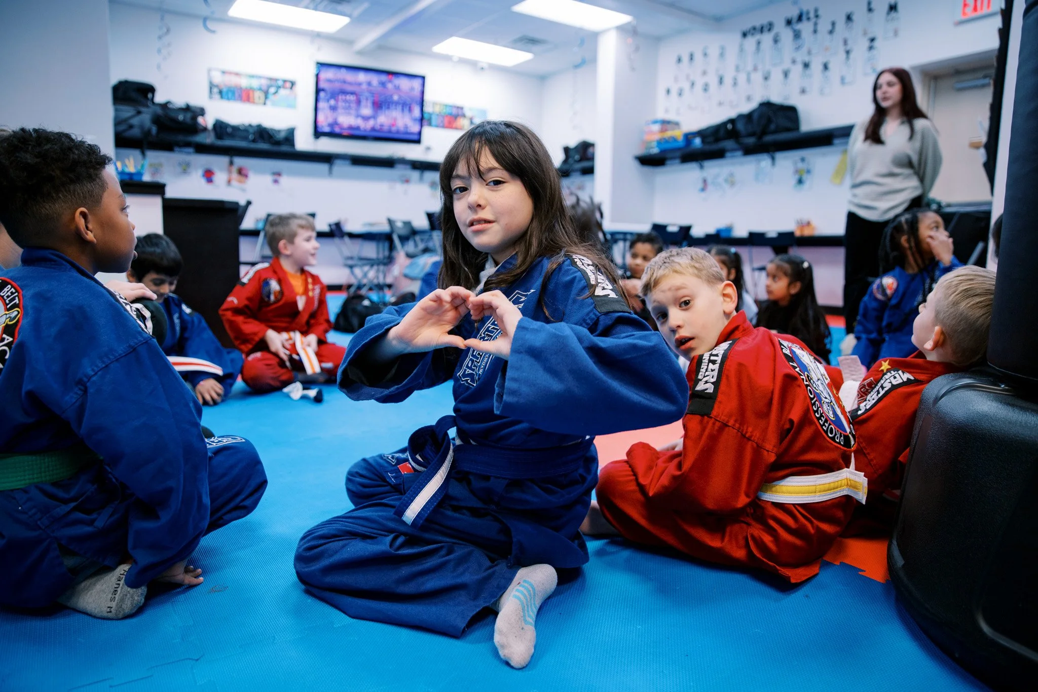 Children in martial arts uniforms sitting on a blue mat in a classroom, one girl in the center making a heart shape with her hands, others around her, with a woman standing in the background.