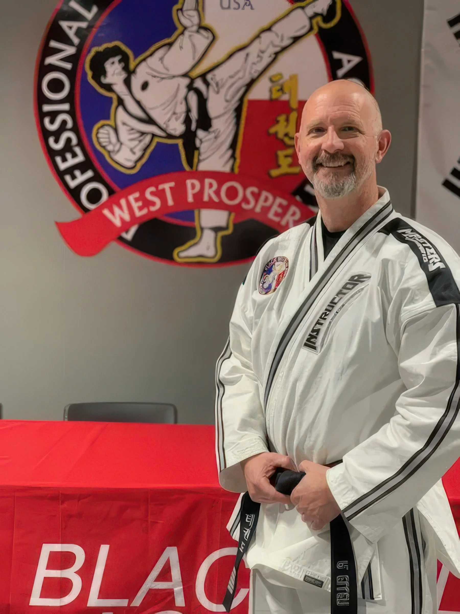 A man in a white martial arts uniform with black stripes, holding a black belt, standing in front of a martial arts banner that reads 'West Prosper' and features a person in a karate pose.