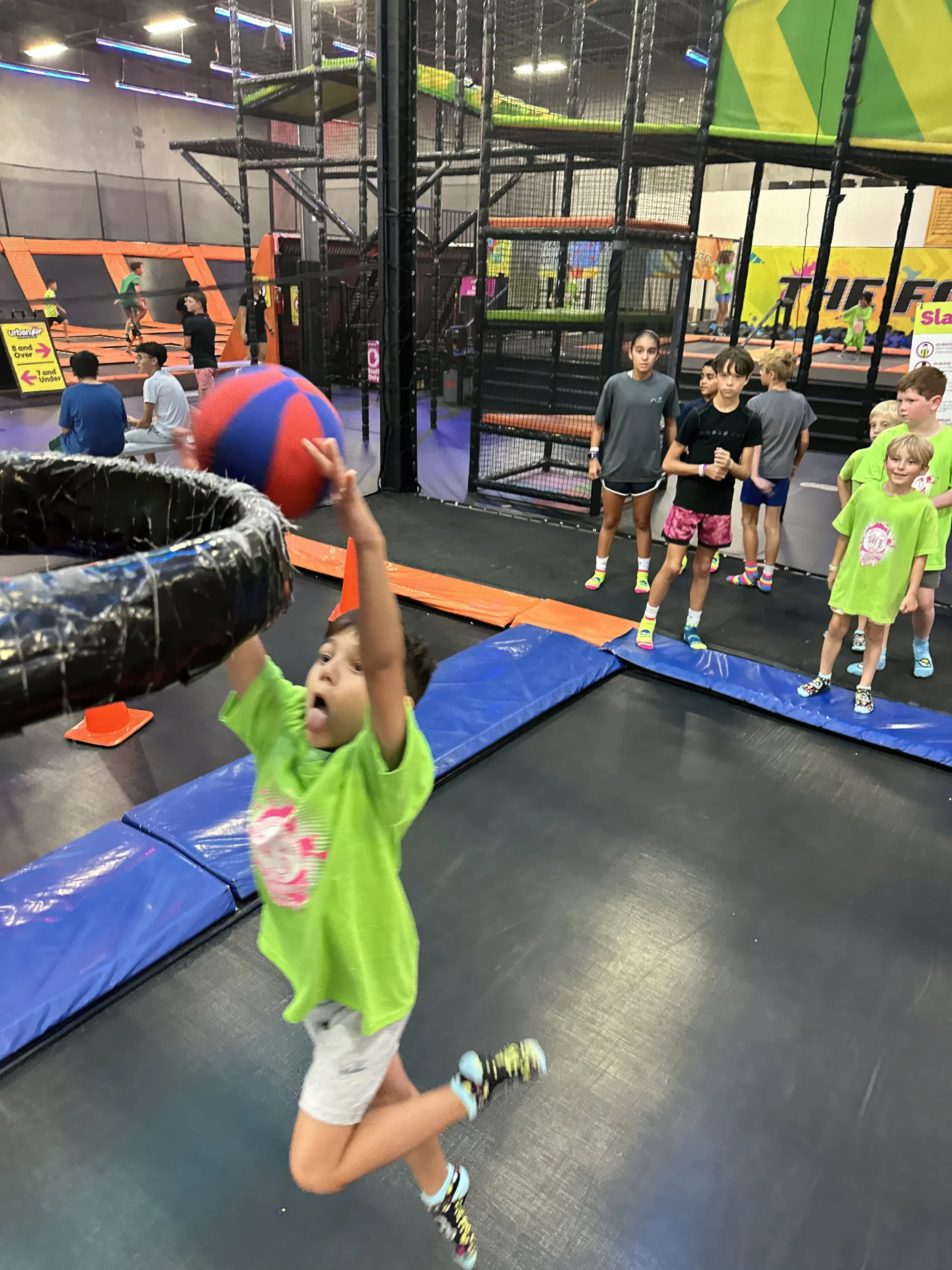 A boy jumping at an indoor trampoline park, reaching for a large red and blue ball, with other children and trampolines in the background.