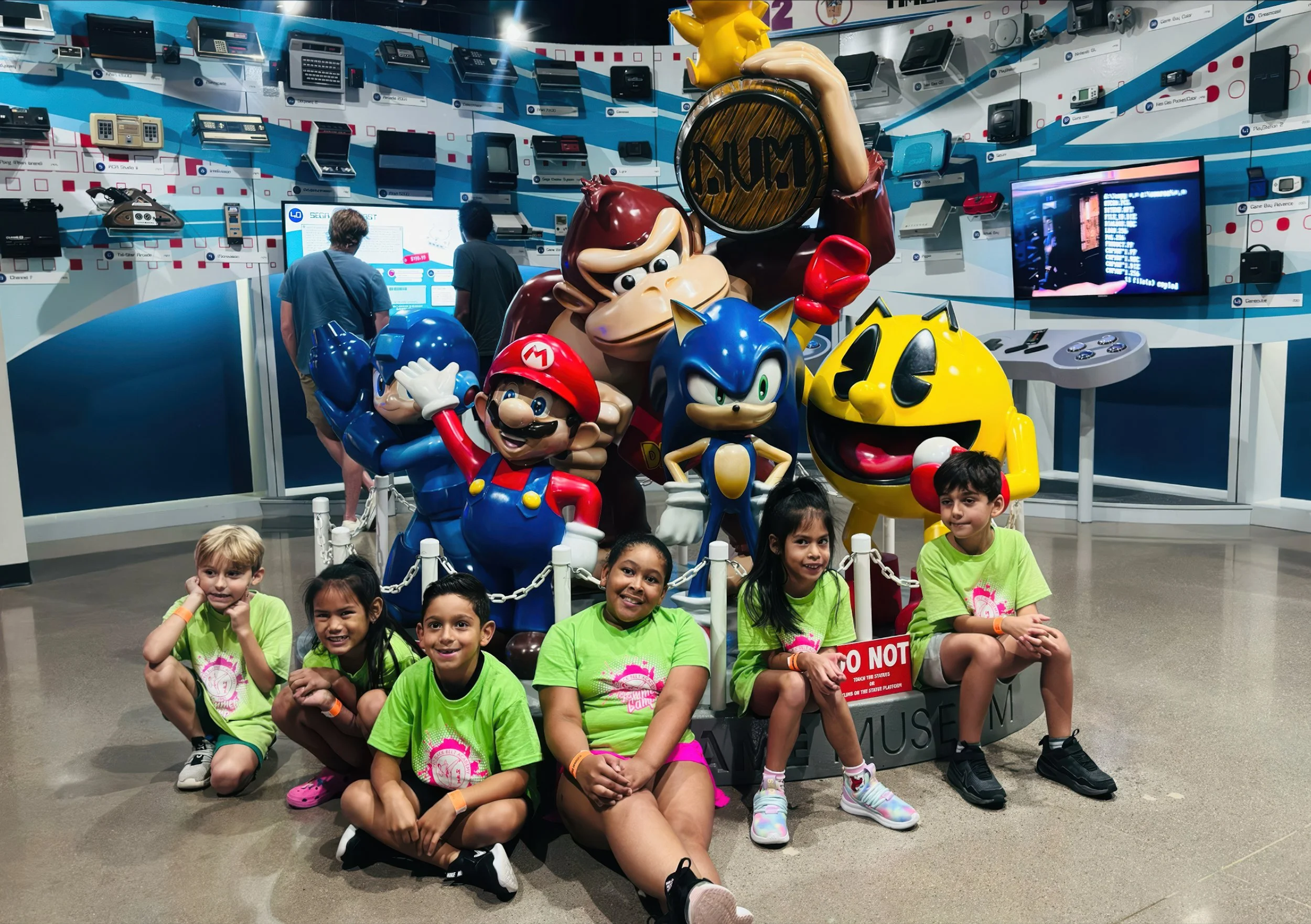 A group of children sitting in front of life-sized figures of popular video game characters Mario, Sonic, Pac-Man, Donkey Kong, and others inside a gaming museum or arcade with a wall of vintage game consoles and a screen displaying game-related cont