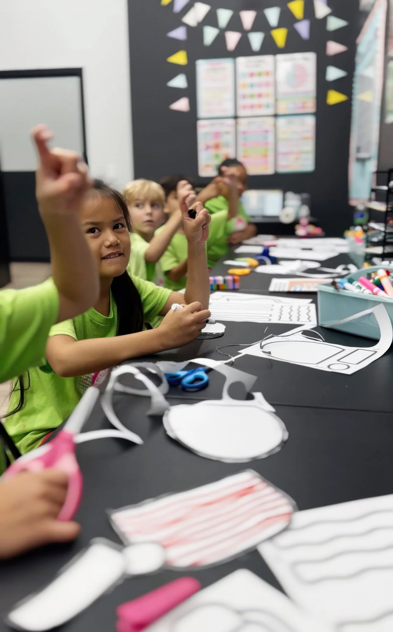 Children in green shirts sit at a table making paper face masks with craft supplies like scissors, markers, and paper. The background features colorful banners and a black wall with educational posters.