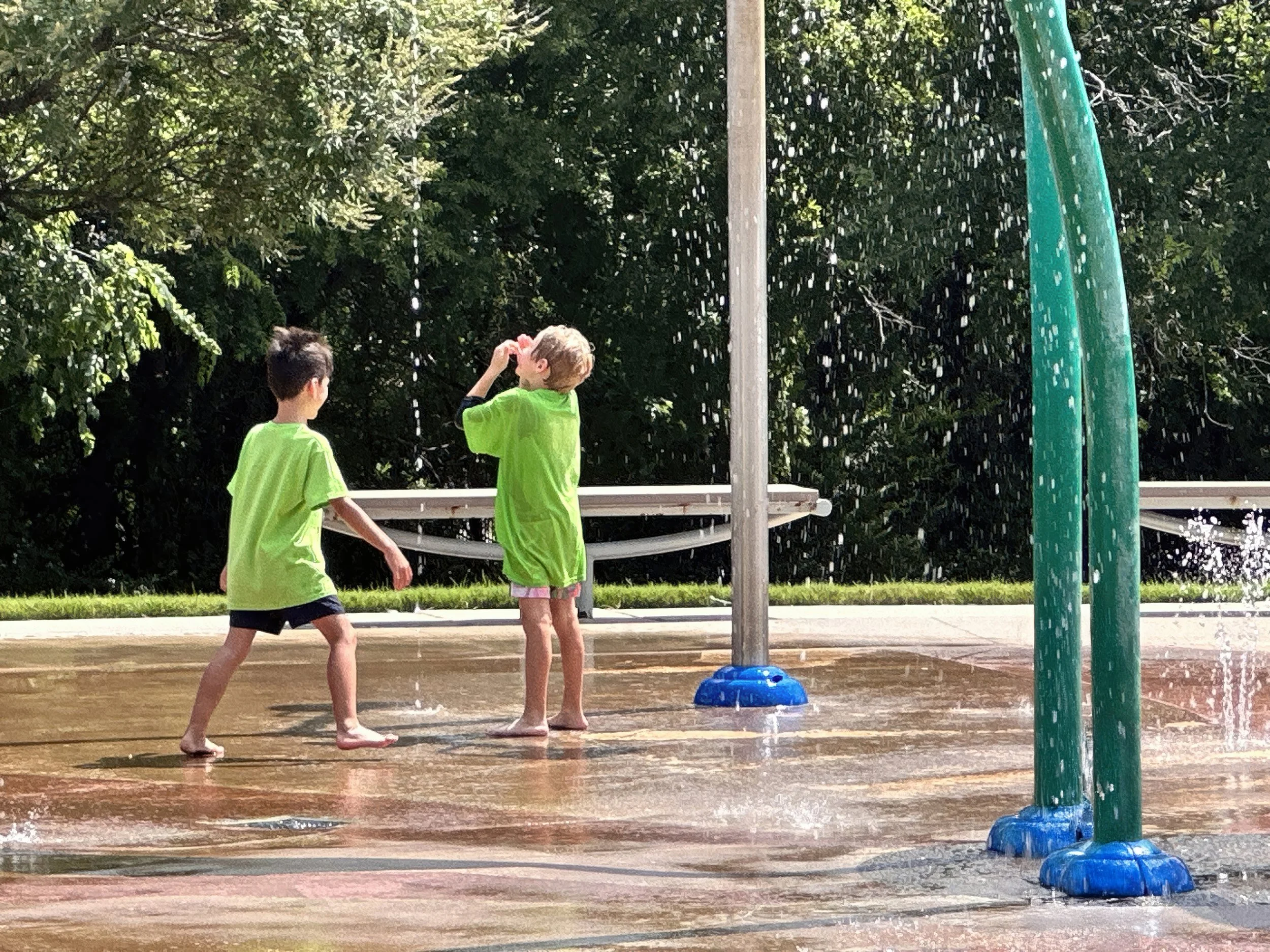 Two children playing at a splash pad in a park, wearing green shirts, with trees in the background.
