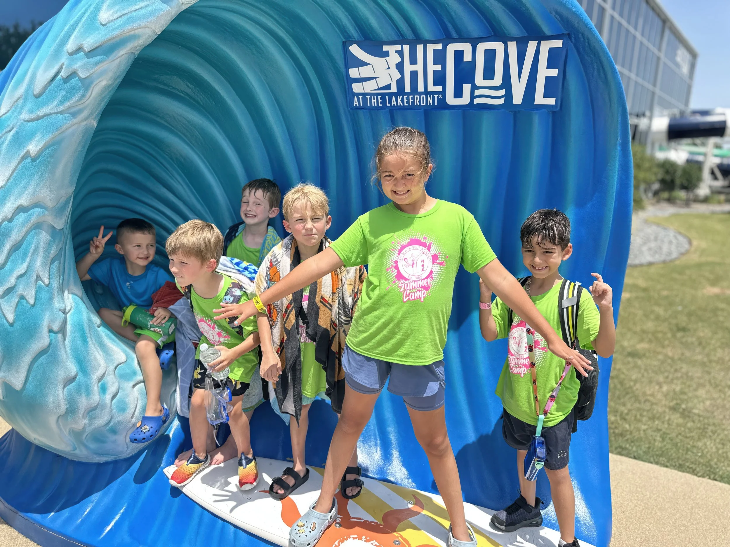 Group of children and a young girl at an outdoor summer camp, standing in front of a large cardboard cutout of a blue wave with the text 'The Cove at the Lakefront.' The children are smiling, some are making peace signs, and they are wearing casual s