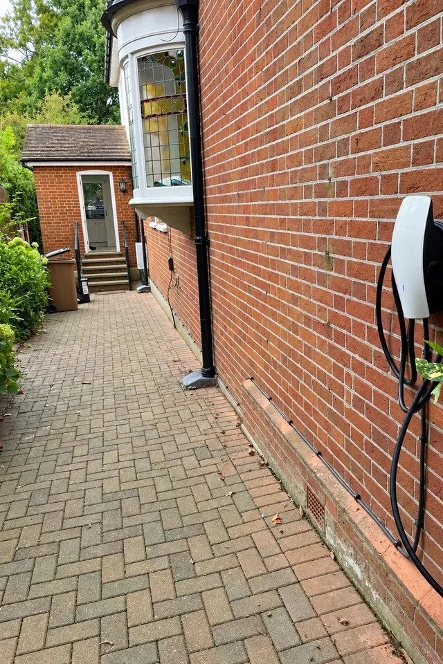 White wall mounted EV charger on a red brick side wall, above a block paved path running past a bay window