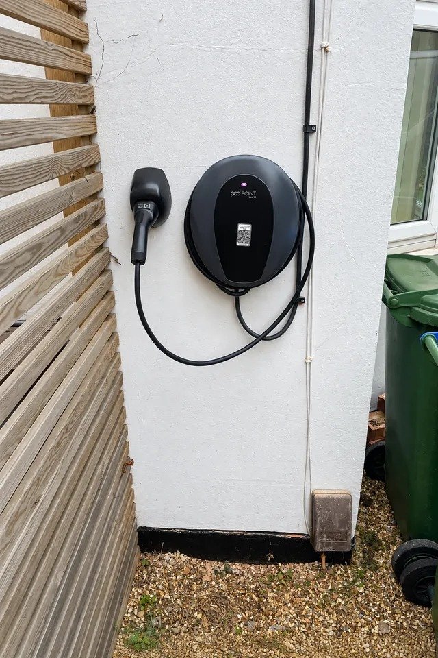 Black wall mounted EV charger with its cable coiled, fitted to a white rendered wall next to a timber fence