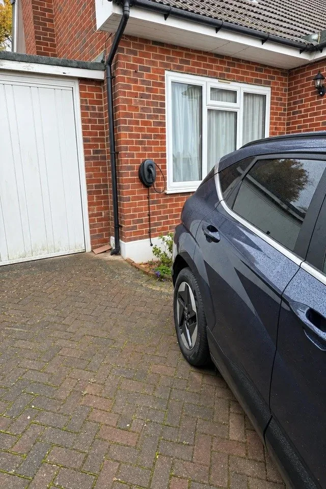 Black wall mounted EV charger on a brick wall beside a white garage door, with a car parked alongside on block paving