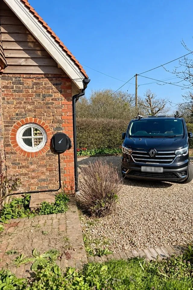 Black wall mounted EV charger on the brick wall of a barn beside a round window, with a cable running down to a van parked on gravel