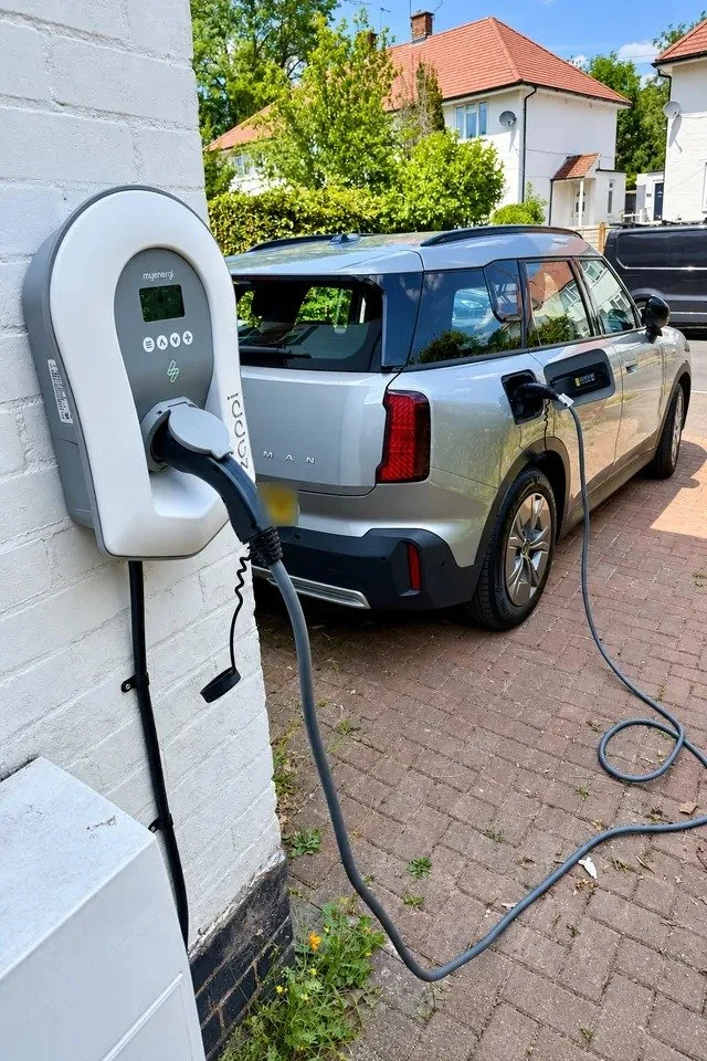 White wall mounted EV charger on a painted brick wall, its cable plugged into a silver car on a block paved driveway