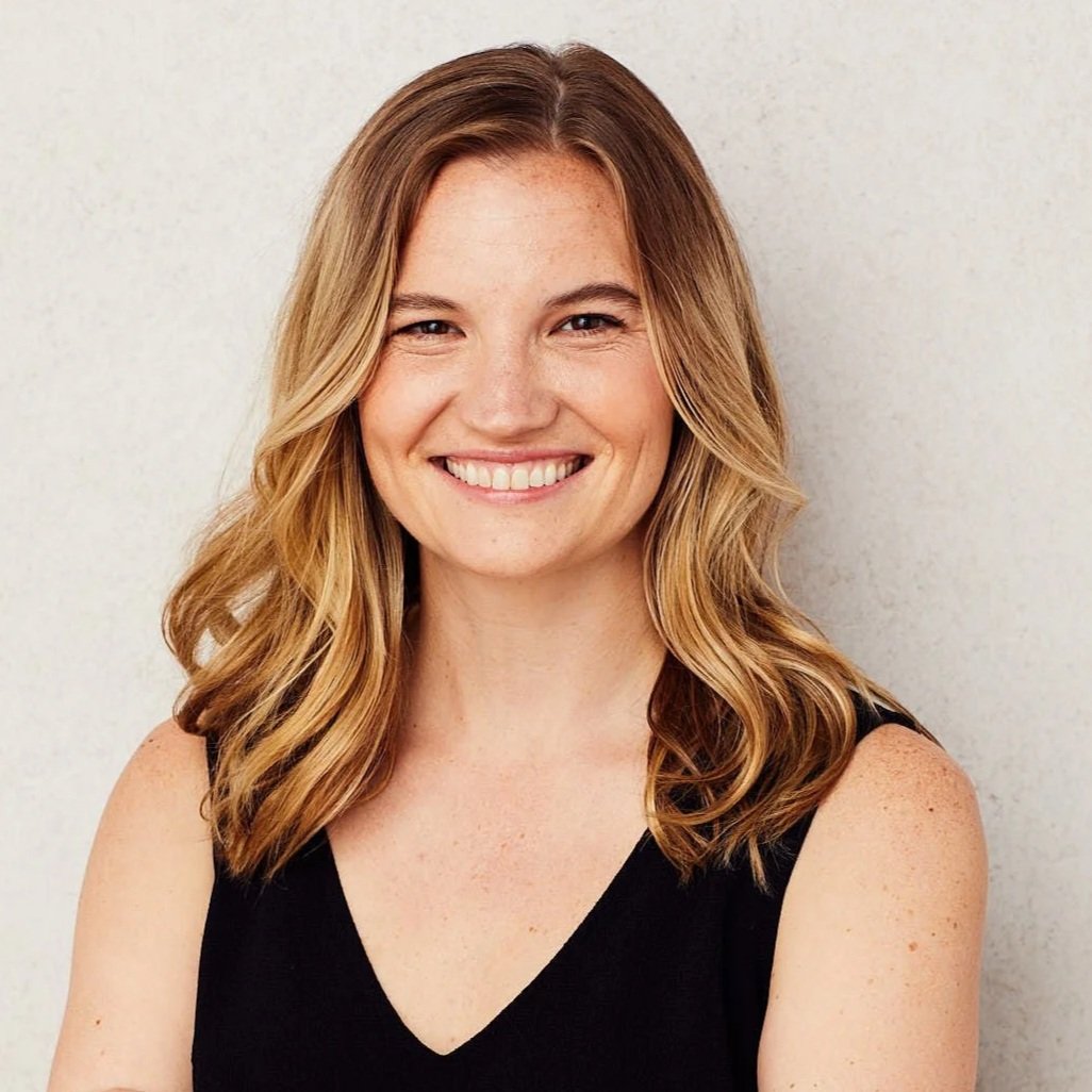 A woman with long wavy blonde hair, laughing and smiling, wearing a colorful scarf and casual clothing, sitting against a plain beige background.