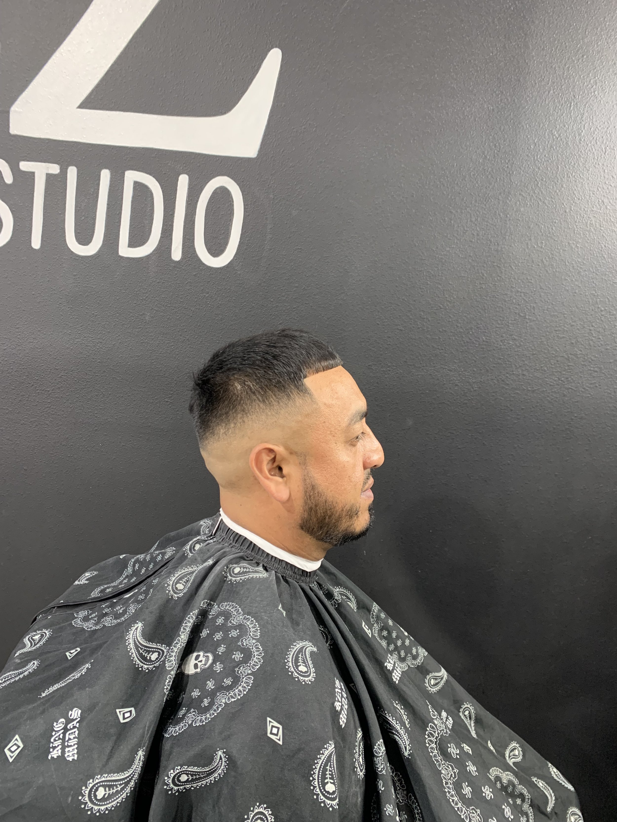 Man with a high fade haircut sitting in a barber shop chair with a black and white bandana cape, in front of a dark gray wall with a logo reading 'STUDIO'.