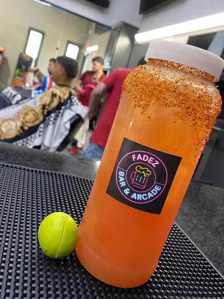 A bottle of orange-colored beverage with a white cap and a black label reading "Fadez Bar & Arcade" with a neon graphic of a beer mug. A lime sits beside the bottle on a black textured surface. In the background, people are sitting at a counter in a
