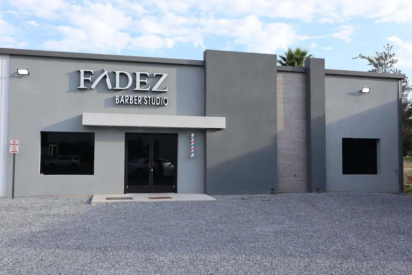 Front view of a modern gray barber shop building with a sign that reads 'FADEZ BARBER STUDIO', a barber pole near the entrance, and a gravel parking area in front.