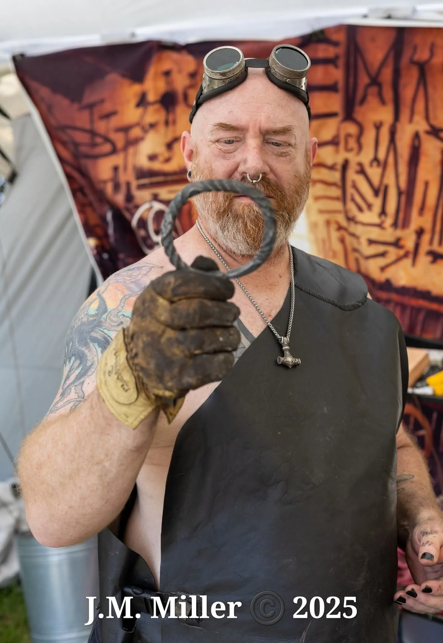 A bearded man wearing goggles on his head, a black apron, and gloves, holding a horseshoe. He has tattoos on his arm and is standing in front of a decorative backdrop with tribal or abstract patterns.