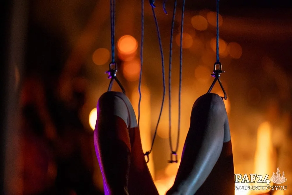 A person hangs from suspension hooks in their knees. A fire is visible in the background.