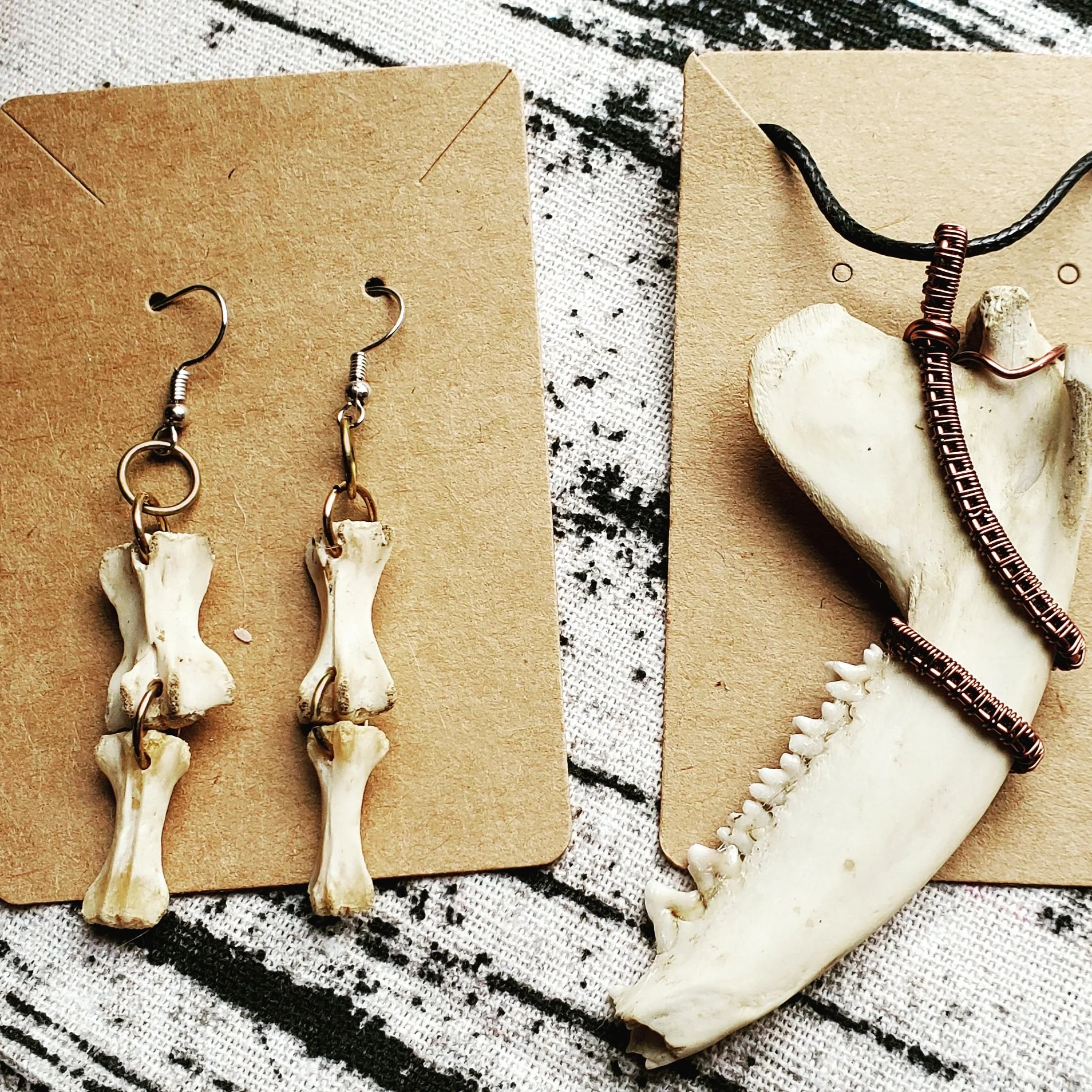 Earrings made from small animal bones and a necklace with a large animal jawbone, adorned with wire wrapping, displayed on brown cardboard backings.