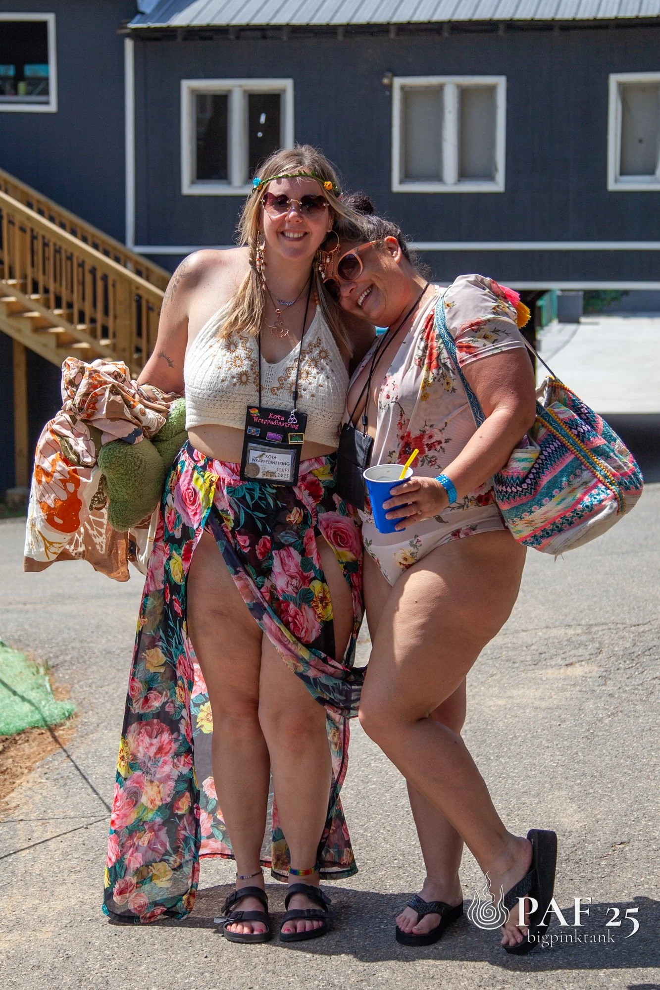 Two women standing close together outdoors, smiling and wearing summer clothing and sunglasses. They have bags over their shoulders and are standing in front of a building with stairs.