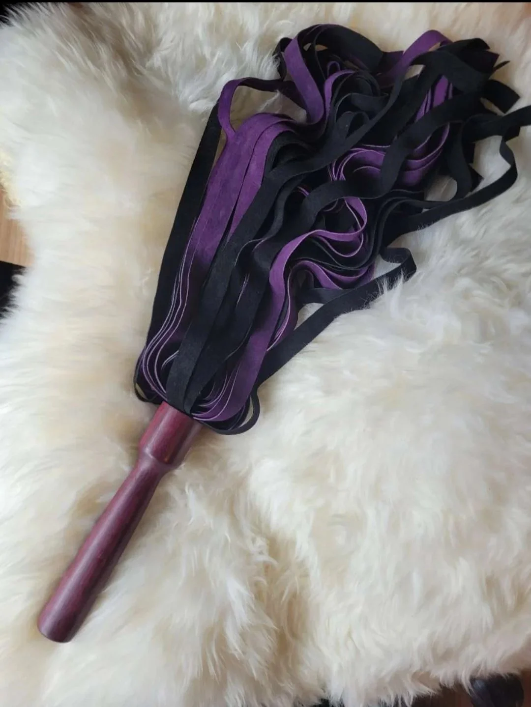 A flogger impact toy with black and purple straps lying on a white furry surface.