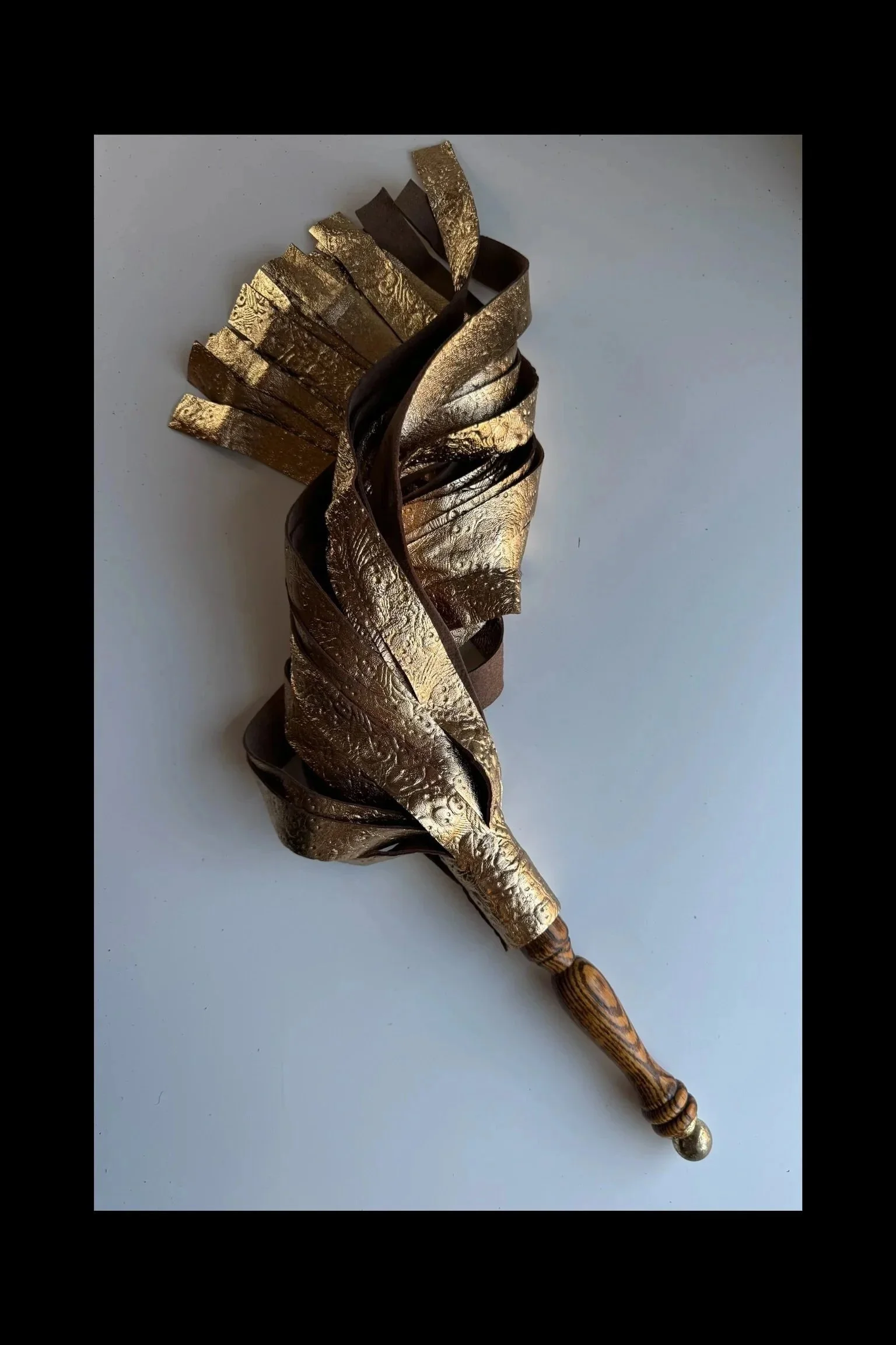 An impact toy resembling a wand with a handle, featuring twisted gold and dark brown or black metallic ribbons or parts around the shaft, with a textured gold surface and a wooden handle, set against a plain background.