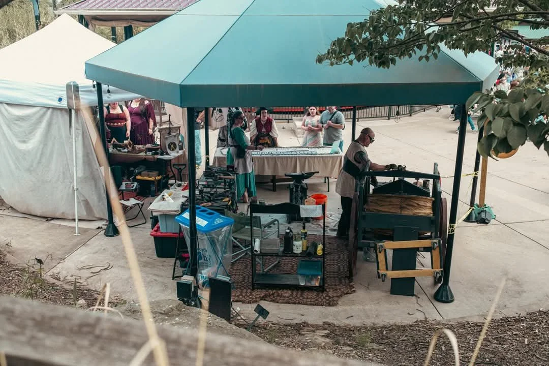 People gathered around a table at an outdoor event, with a person working on a blacksmithing or metalworking project under a teal canopy. There are tents and other people in the background.