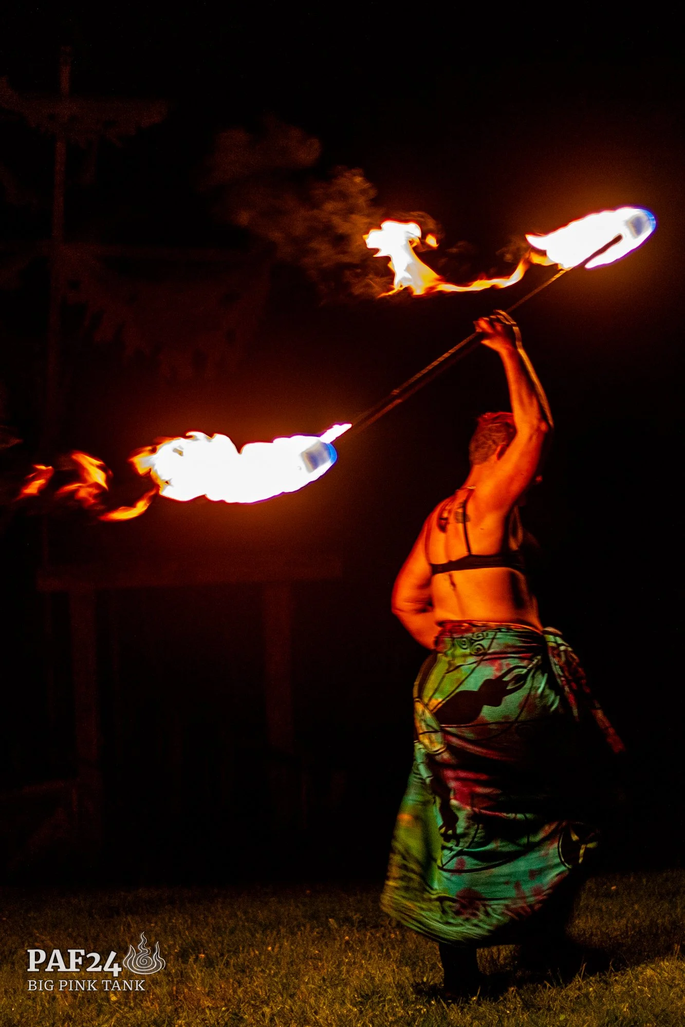 A performer is spinning fire poi at night, creating flames with fire on both ends of a chain, with a dark background and some trees visible.