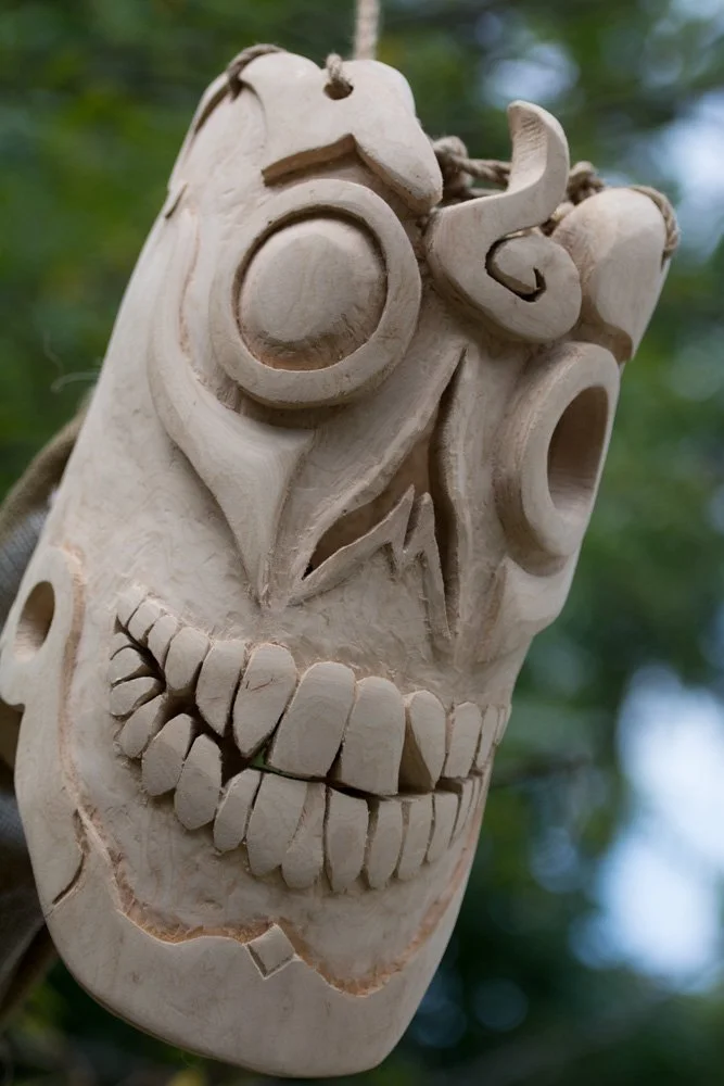Wooden carved skull with exaggerated facial features and open mouth, hanging outdoors with blurred green background.