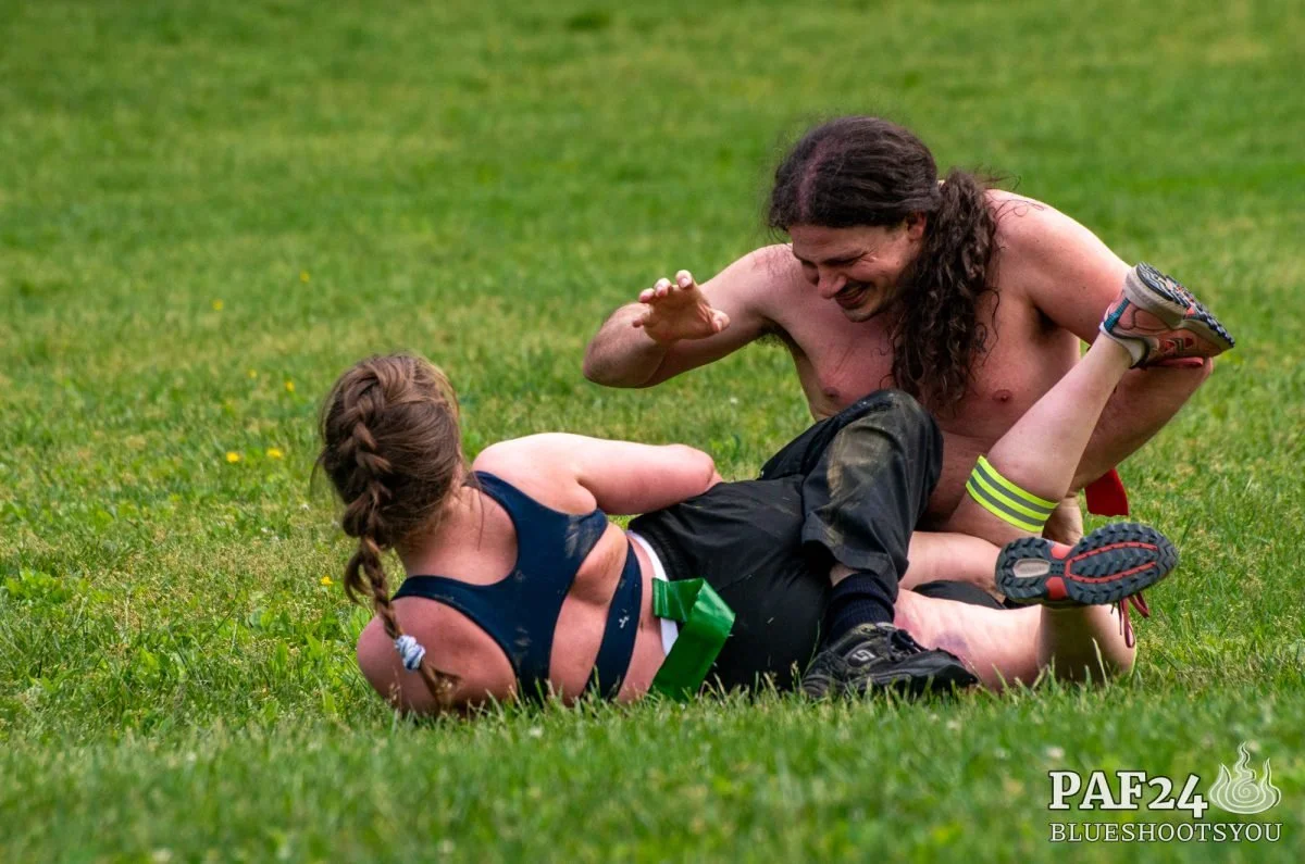 Two people with long hair wrestling on a grassy field, one appears to be a male and the other a female, both shirtless and wearing athletic shorts.