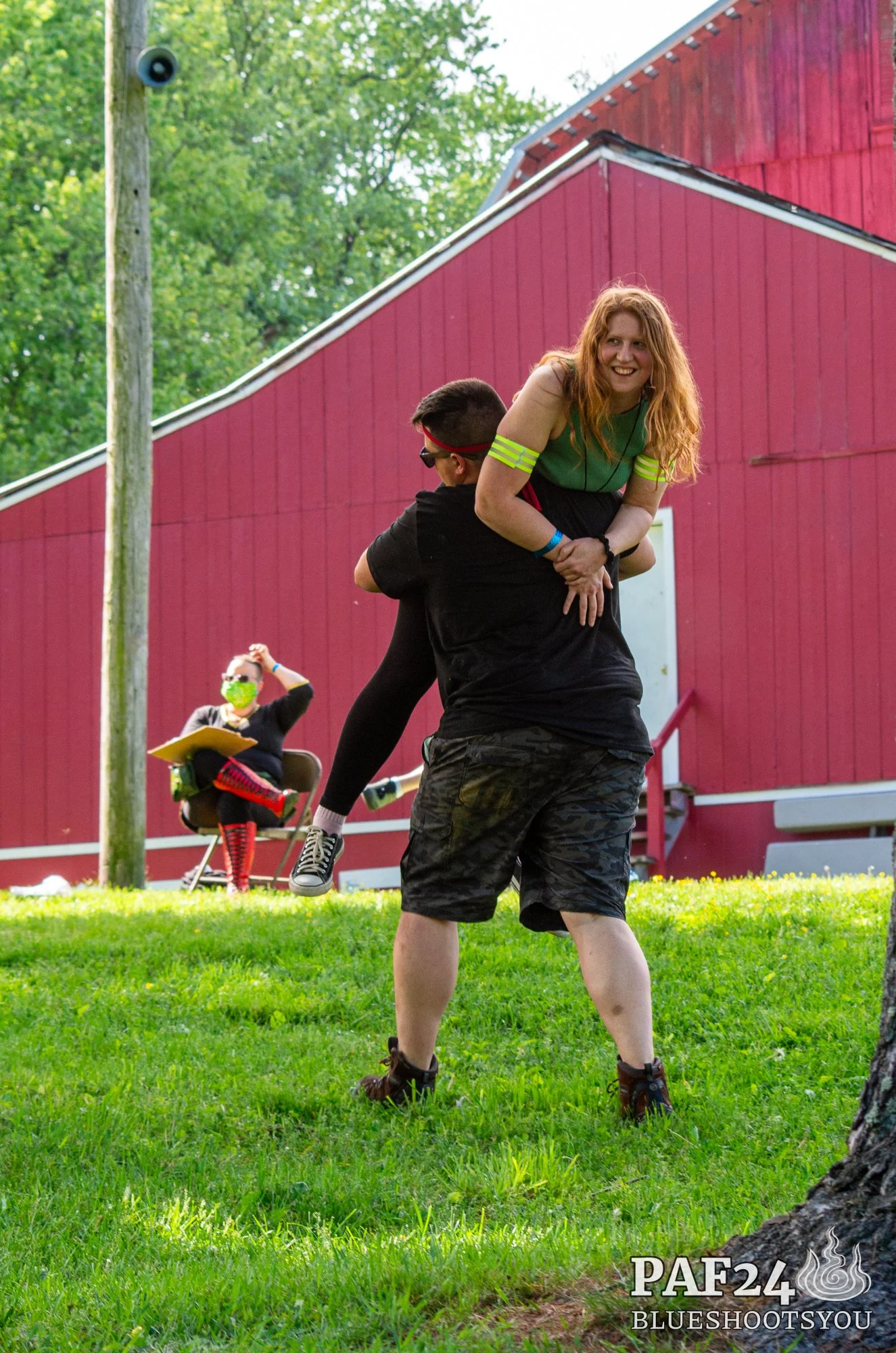 A man giving a shoulder ride to a smiling woman with curly hair, wearing a green shirt with yellow stripes on the sleeves, outside on a grassy area near a red barn, with a person sitting at a table in the background wearing a mask and sunglasses.