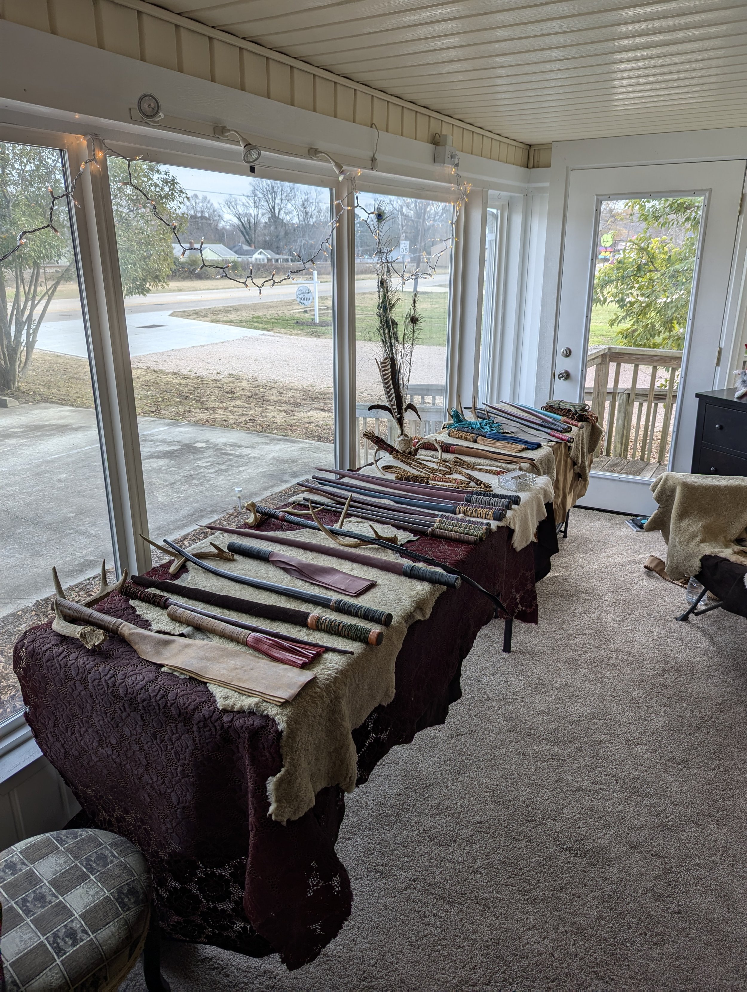 Table displaying various traditional bows and arrow quivers inside a sunroom with glass windows, carpeted floor, and a door leading outside.
