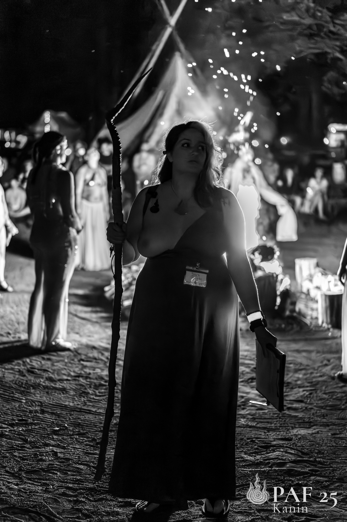 A woman with long hair holding a staff and a clipboard standing at night at a festival or outdoor event, with other people and tents in the background, illuminated by string lights.