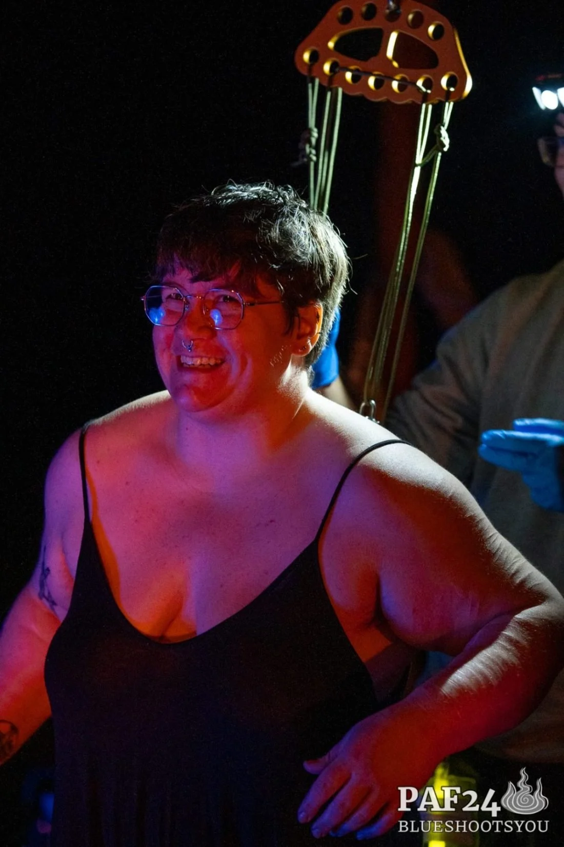 A smiling person with short dark hair, glasses, and a septum piercing, wearing a black spaghetti strap top. Their back is attached to a suspension rig. They are illuminated by colorful stage or party lighting, with a person in the background.