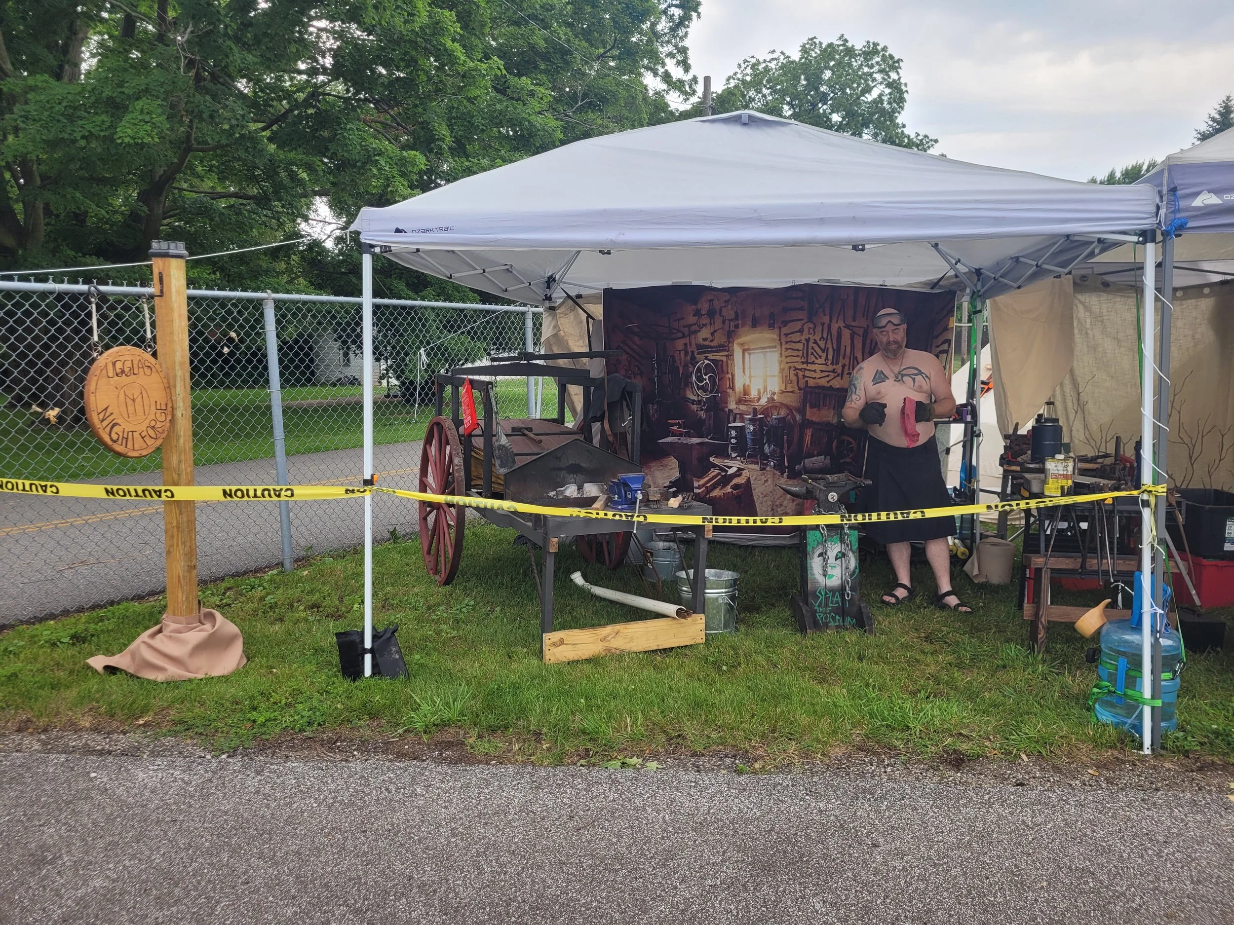 A man standing inside a metal tool and art workshop tent on a grassy area, working on a project with tools, surrounded by workshop supplies, with a backdrop of a fictional workshop scene.