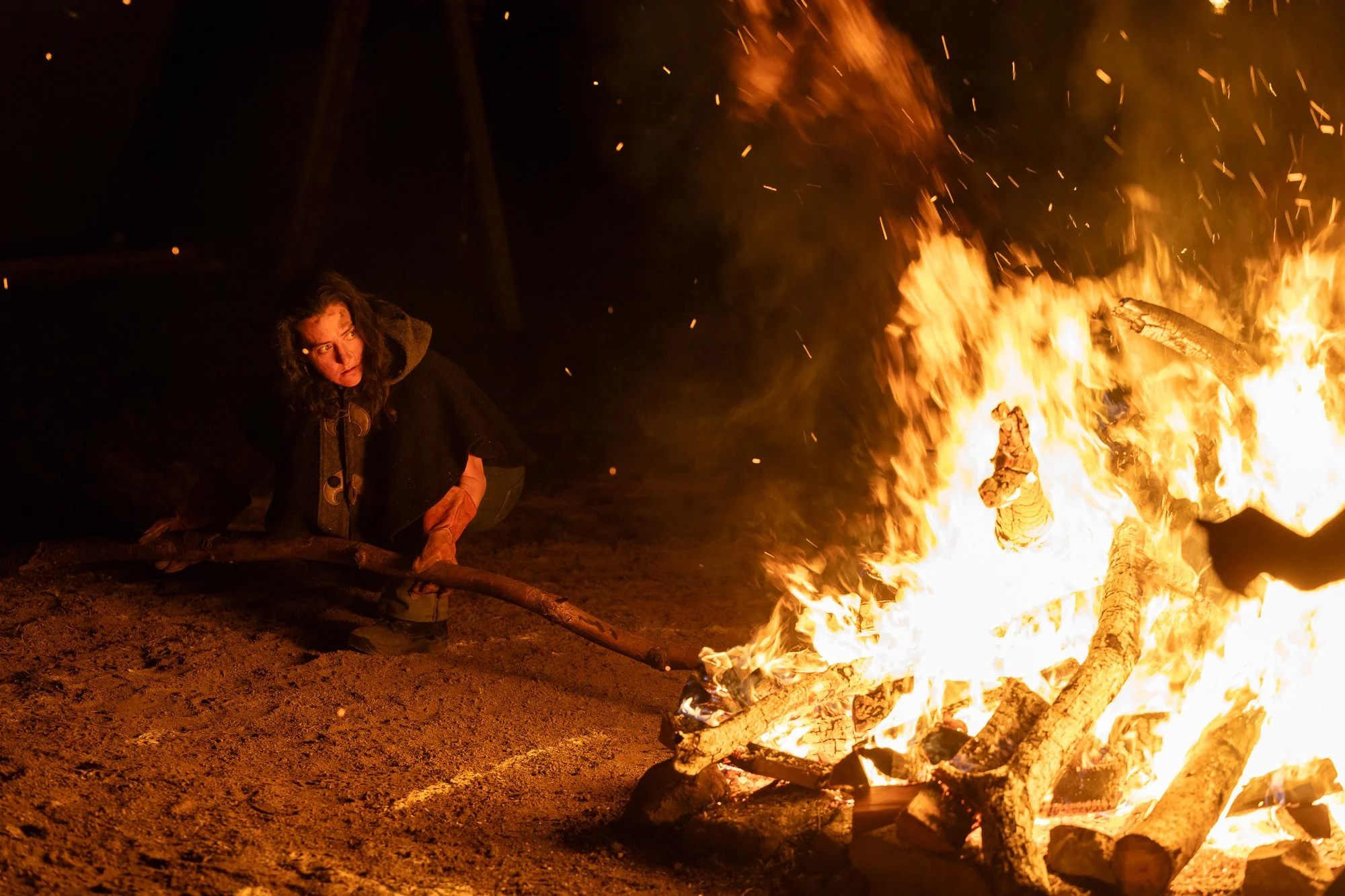 A woman with curly hair wearing a dark cloak is crouching and tending a campfire with large logs burning, sparks flying, in the dark night.