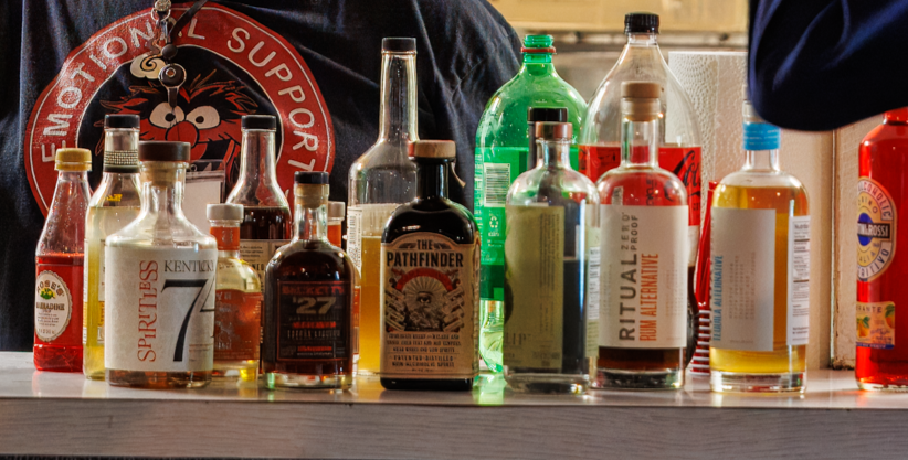 A variety of colorful liquor bottles on a bar counter with two people partially visible behind, one wearing a shirt with a large red badge.
