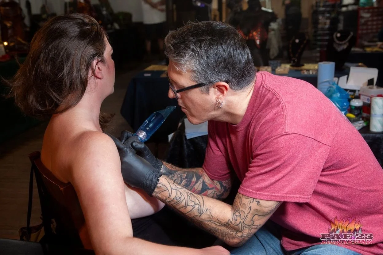 Tattoo artist with glasses and tattoos on arms giving a piercing to a woman with short brown hair, in a tattoo studio with tables and supplies in the background.