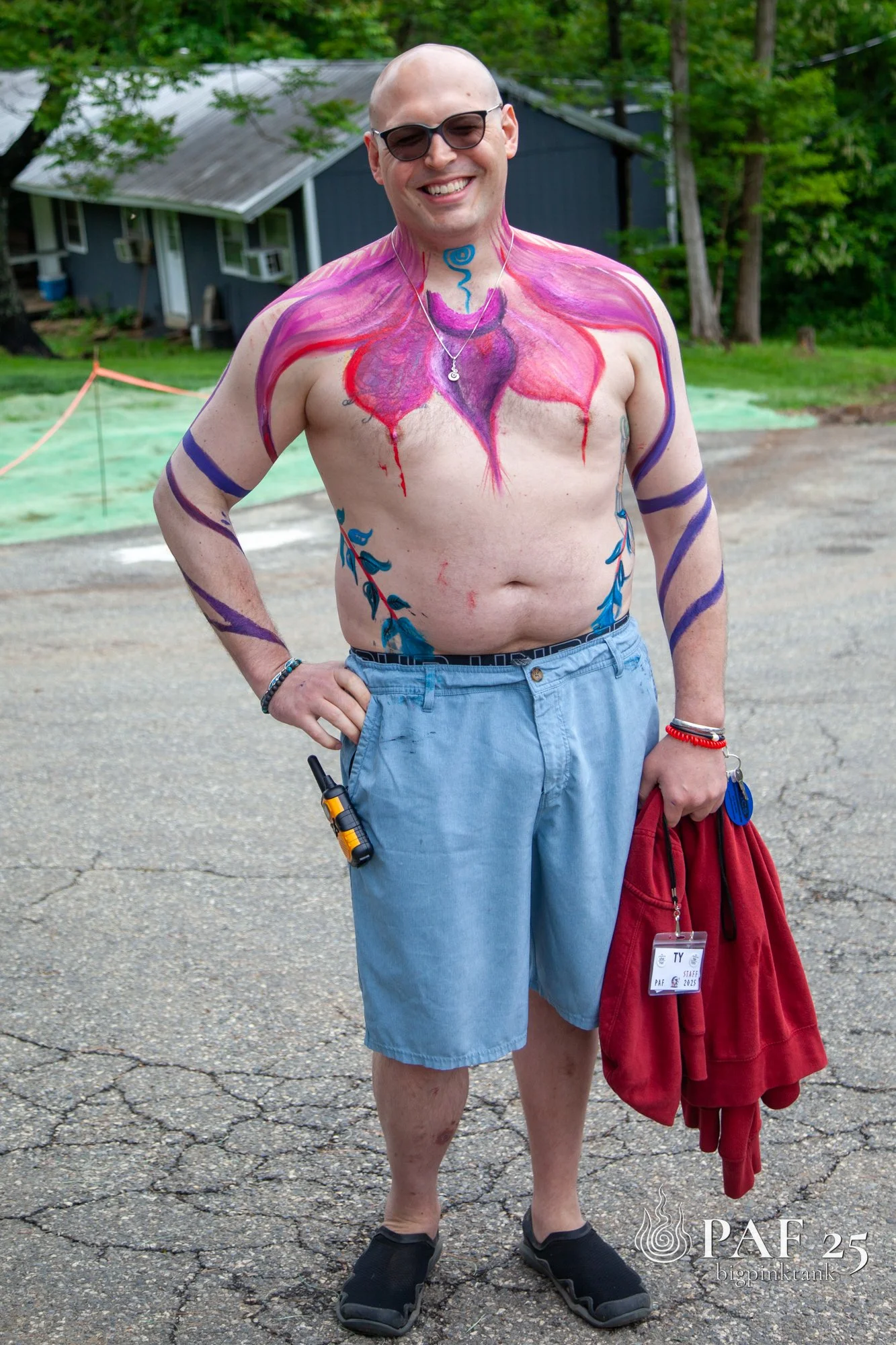 A shirtless person standing outdoors on a paved surface, smiling, with colorful body paint resembling a heart and vines on his chest and arms, holding a red sweatshirt and wearing sunglasses and shorts.